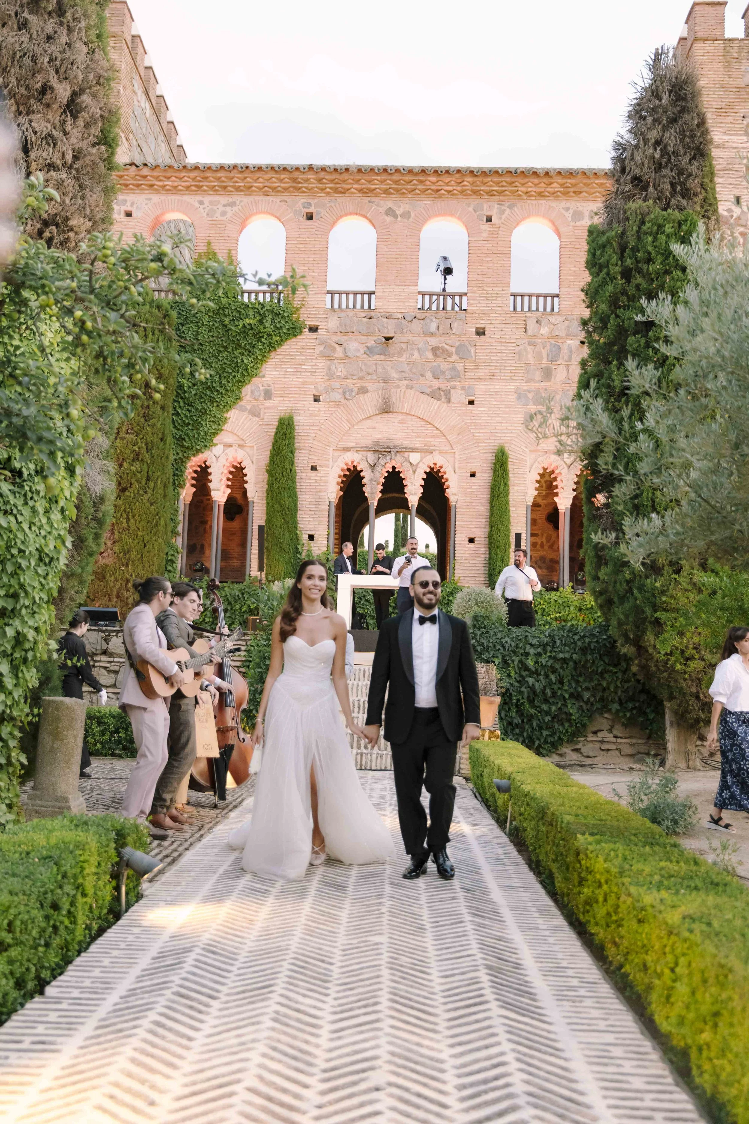 Pareja de recién casados caminando en un pasillo de mármol, rodeados de árboles y músicos tocando el piano y la guitarra, en una ceremonia en un lugar histórico de arquitectura rústica.