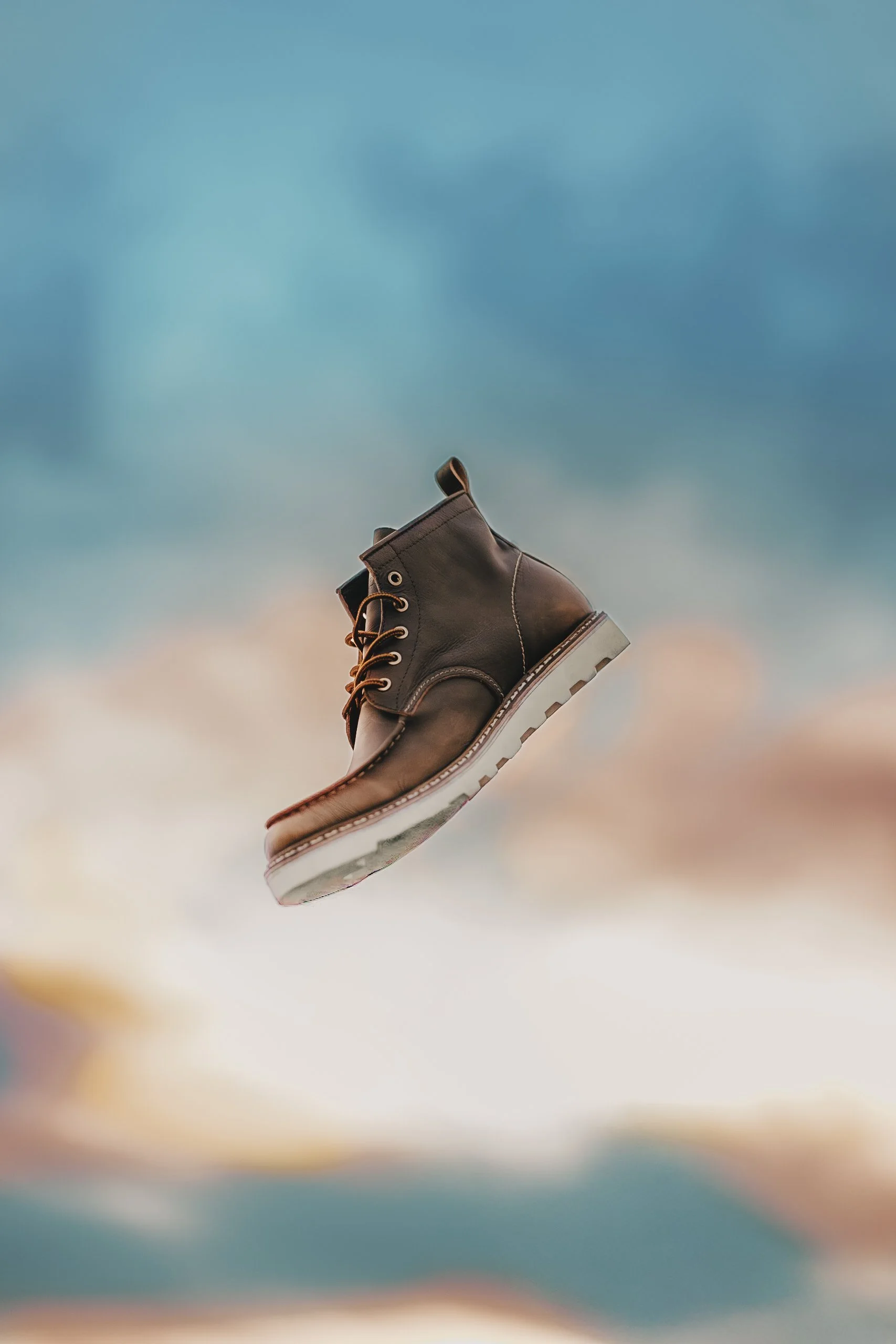 Botín de cuero marrón con suela gruesa, suspendido en el aire contra un cielo con nubes y tonos de atardecer.