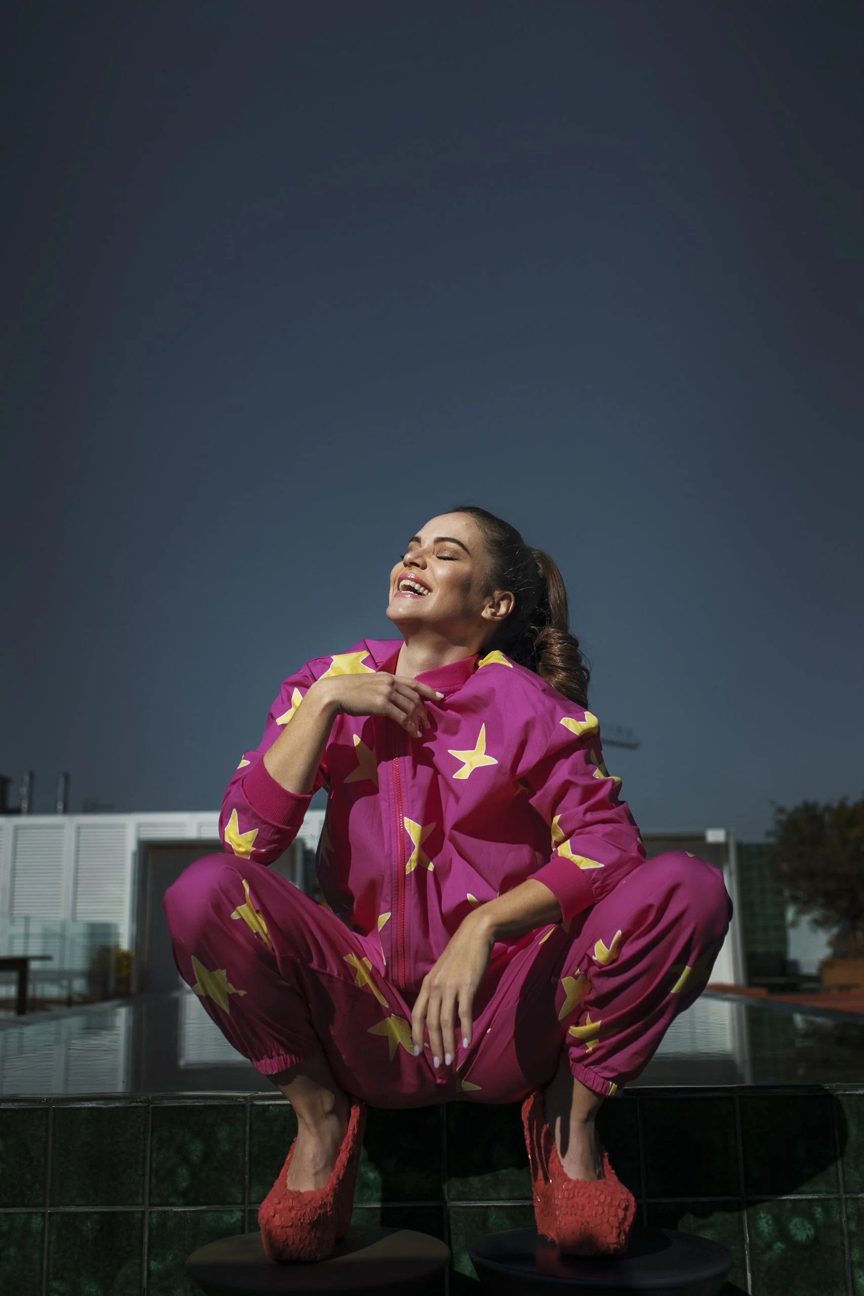 Una mujer sonriente con ropa deportiva rosa con estrellas amarillas, descansando sobre bloques en un espacio abierto en la noche.