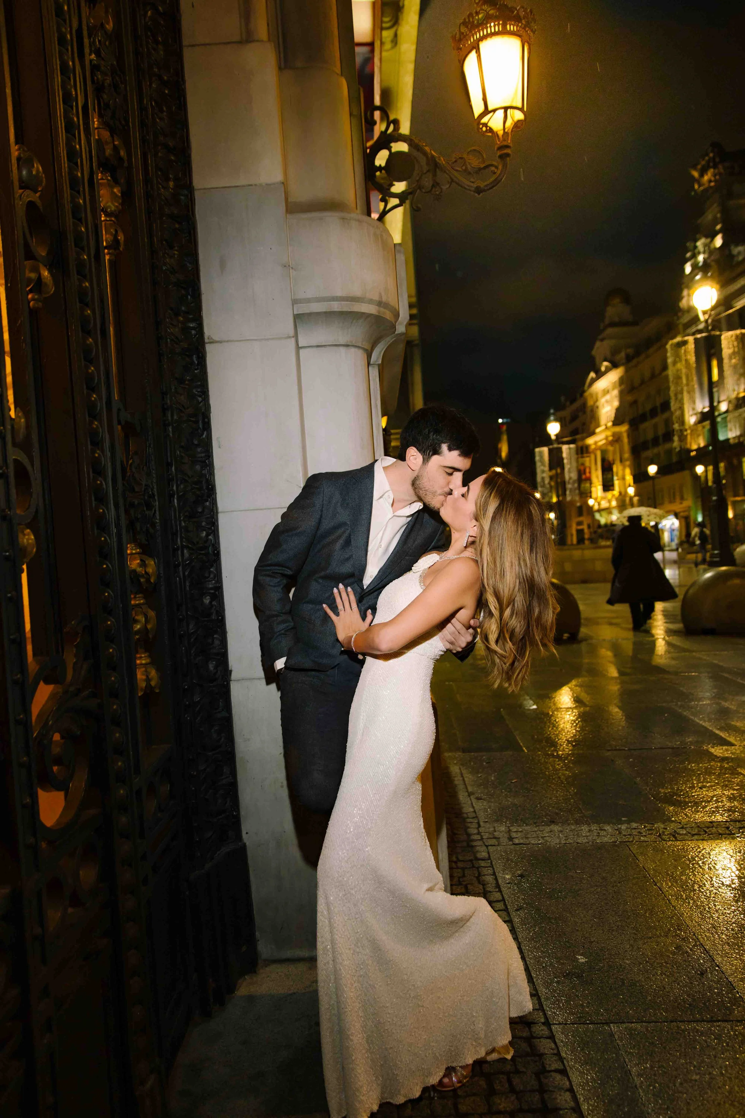 Una pareja besándose en la calle de noche, con la mujer en un vestido largo y el hombre en un traje, junto a una entrada con una puerta de hierro decorada, y faroles que iluminan la escena.