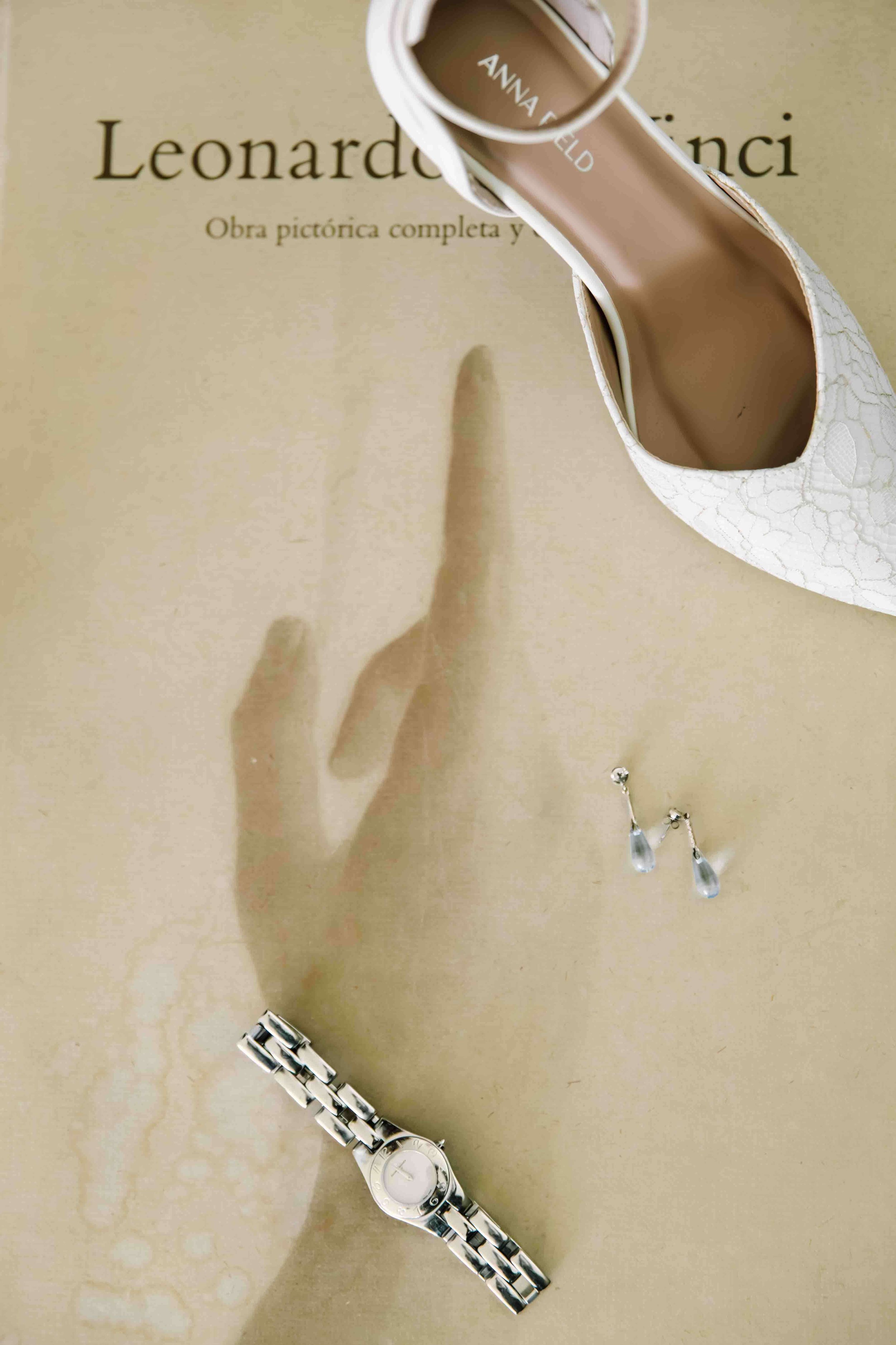 Un zapato de tacón blanco con detalles de encaje, un reloj de pulsera de metal, unos aretes con gotas de cristal y un libro de Leonardo da Vinci en fondo beige.
