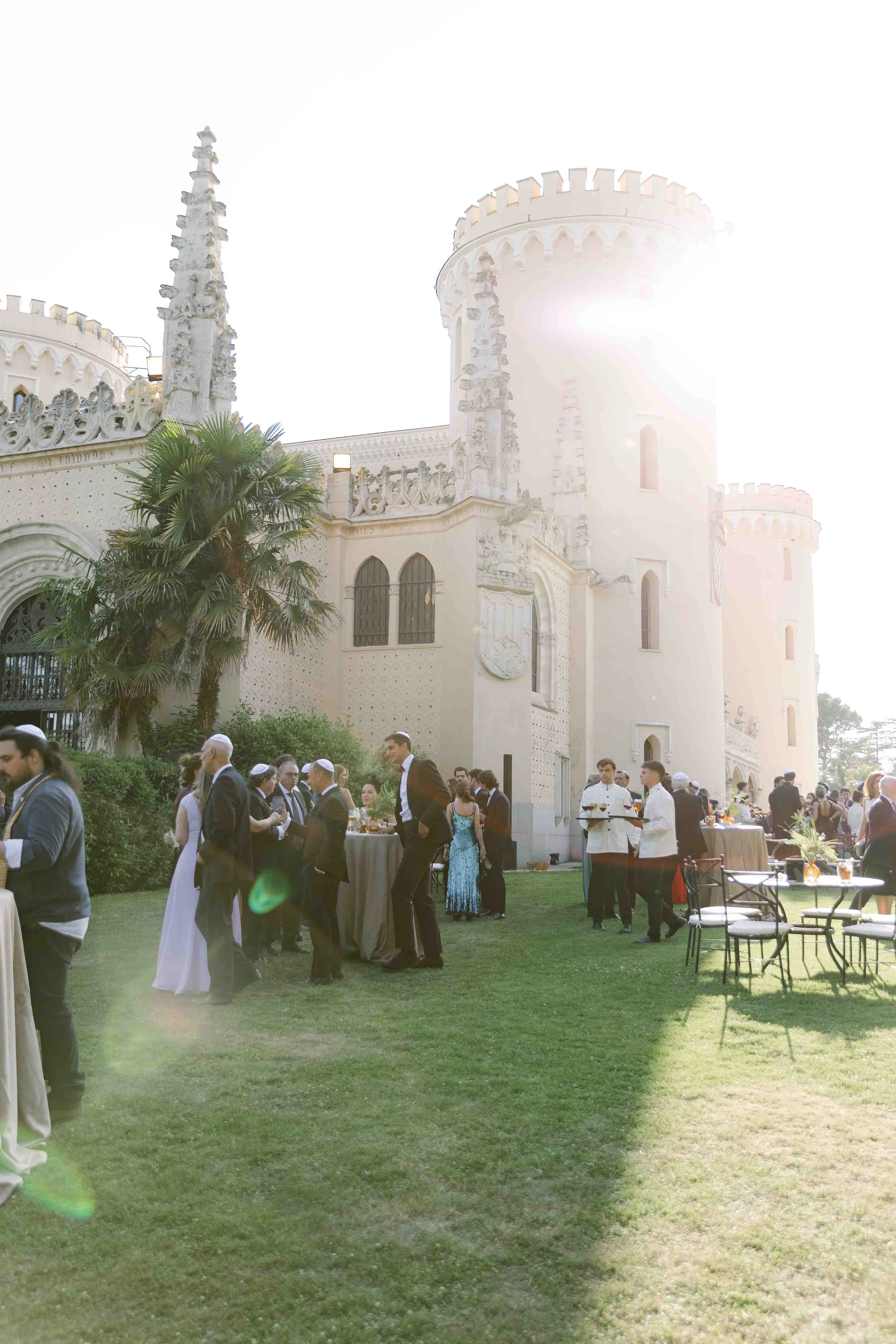 Celebración al aire libre en los jardines del castillo, con invitados vestidos formalmente, mesas con comida y bebida, y la estructura del castillo visible en el fondo.