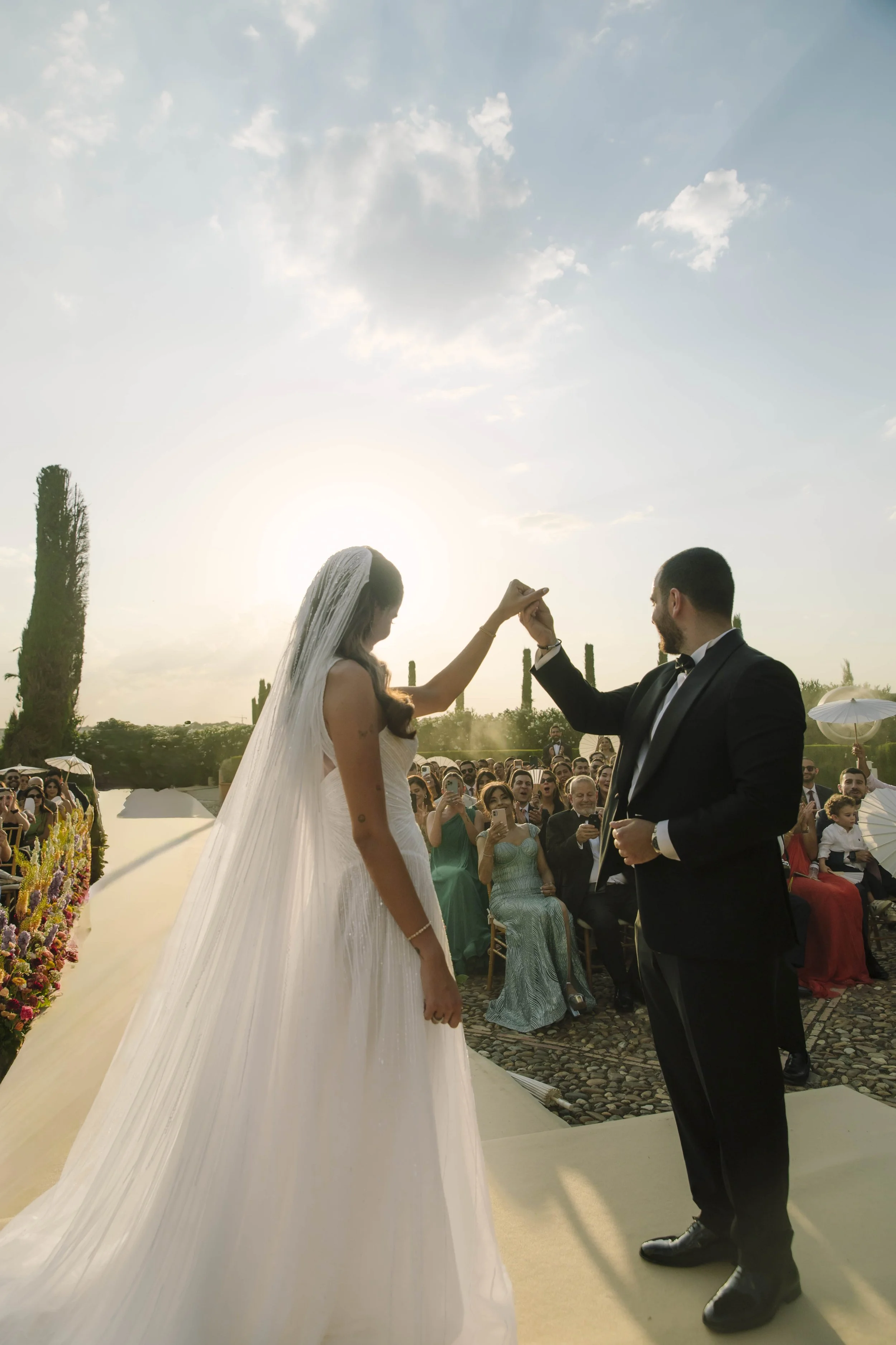 Pareja de novios en boda al aire libre, el novio en esmoquin negro y la novia con vestido blanco y velo, durante la ceremonia, con invitados observando y tomando fotos, bajo un cielo soleado.