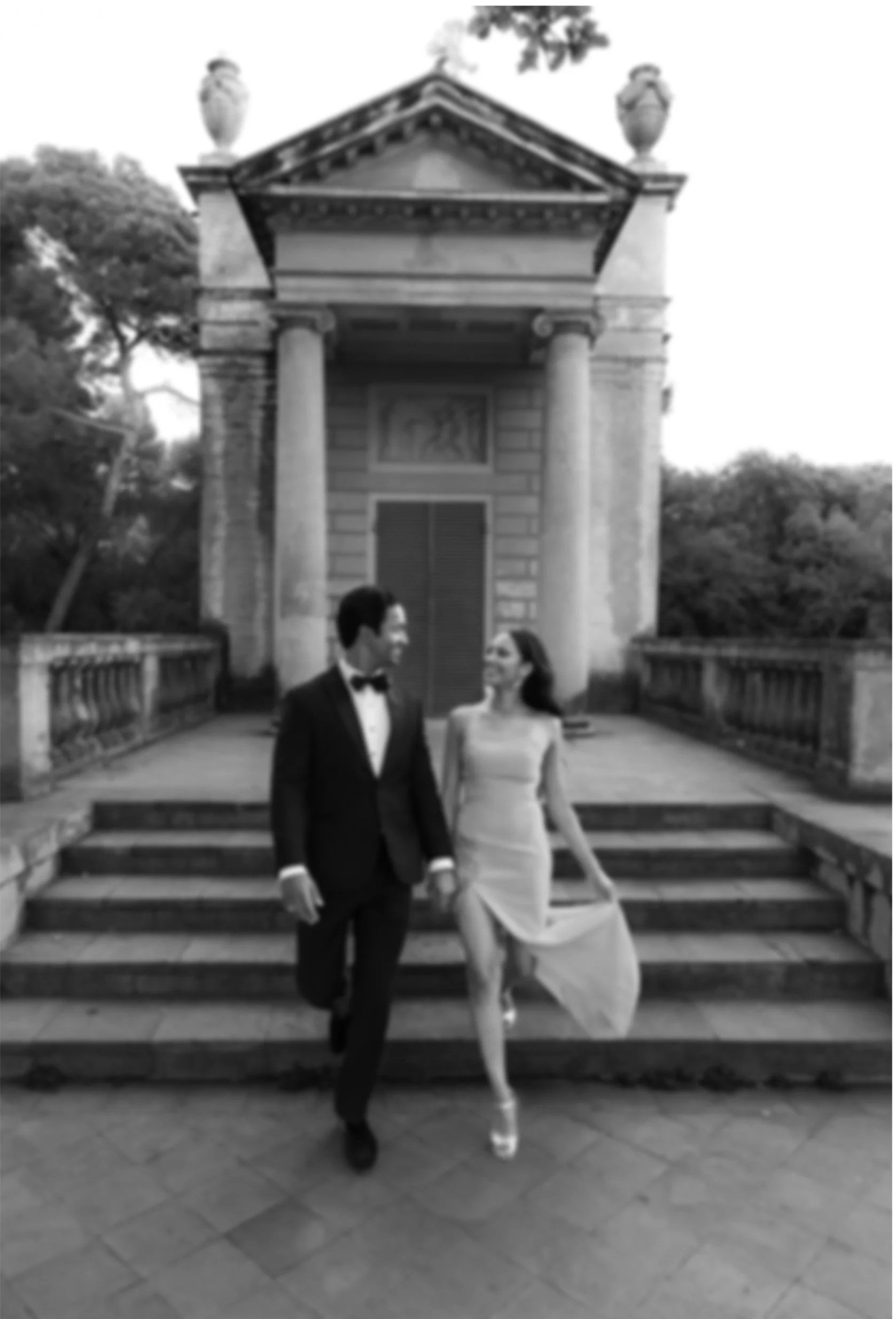 Pareja vestida con ropa elegante caminando por unas escaleras frente a una estructura arquitectónica antigua, en un entorno al aire libre, en blanco y negro.