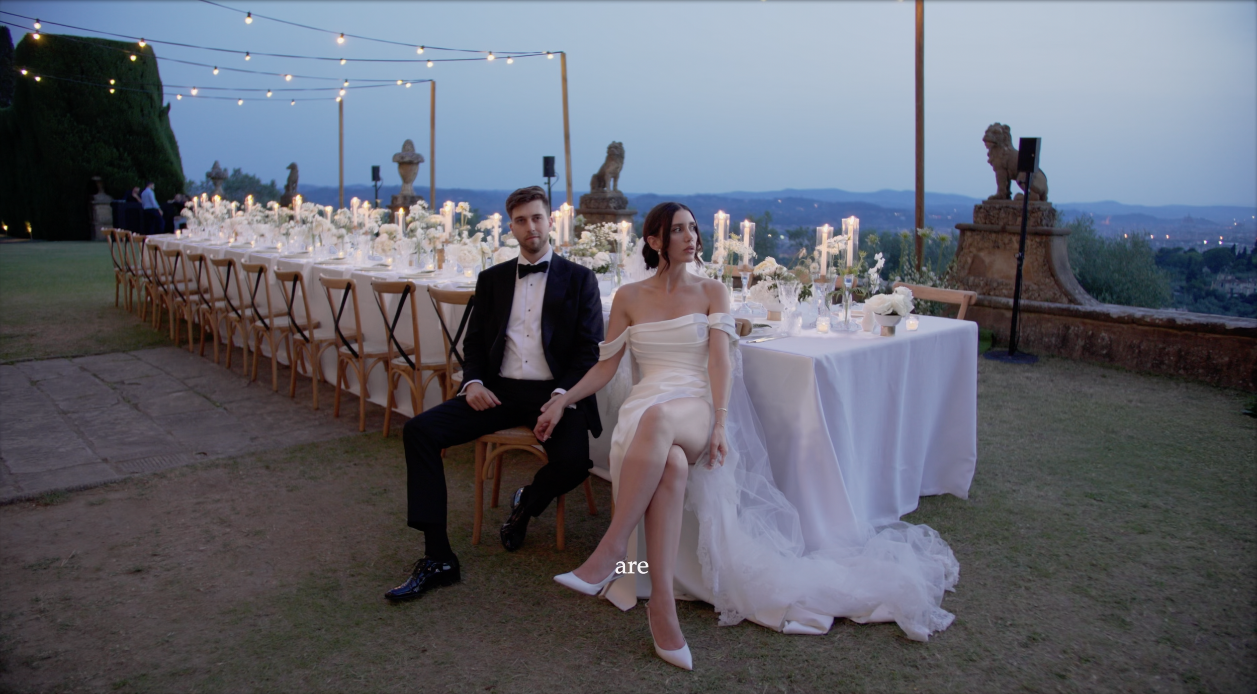 Pareja en vestido de novia y traje de novio sentados en la mesa de una boda al aire libre con decoración elegante y velas, en un lugar con vista panorámica y luces colgantes.