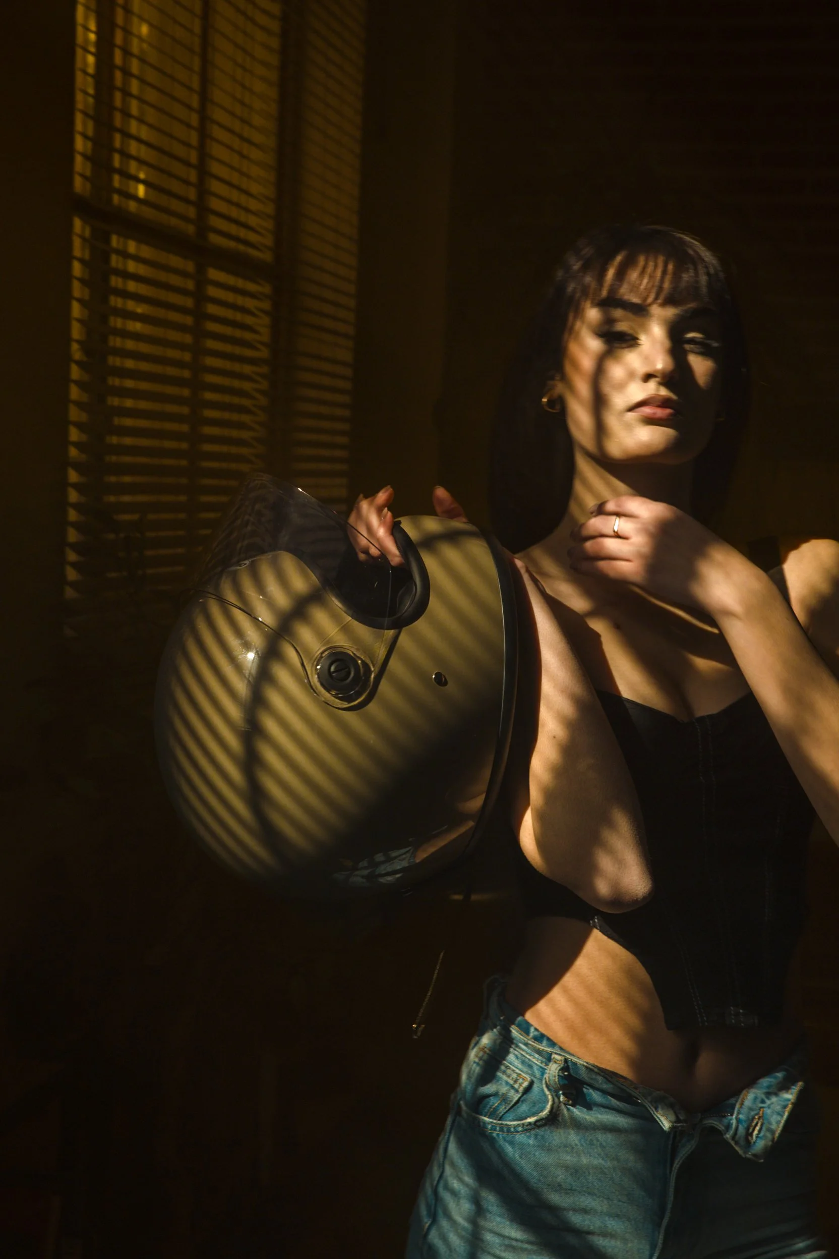 Mujer con cabello oscuro y flequillo, sosteniendo un casco de motocicleta en la mano, en un ambiente con luz y sombras desde persianas. Ella lleva una camiseta sin mangas negra y pantalones vaqueros.