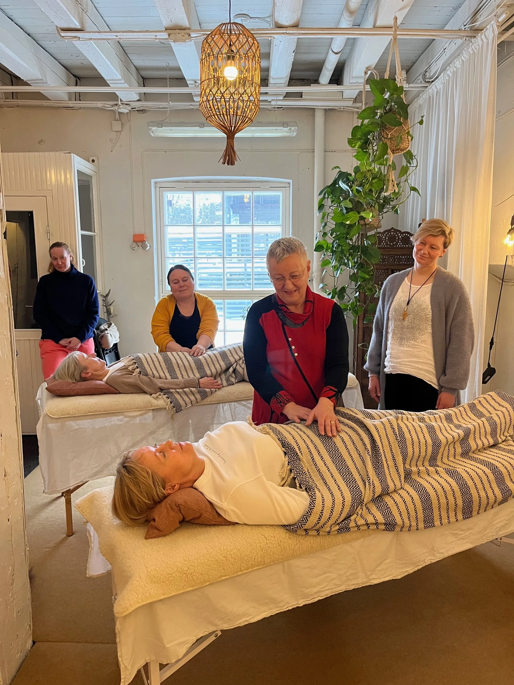 Kuusi naista ja kaksi naispientä istuu ja makaa, osa on makuupusseissa, huoneessa jossa on suuri ikkuna ja koristeita, jotka osallistuvat todennäköisesti jooga- tai rentoutustilaisuuteen.