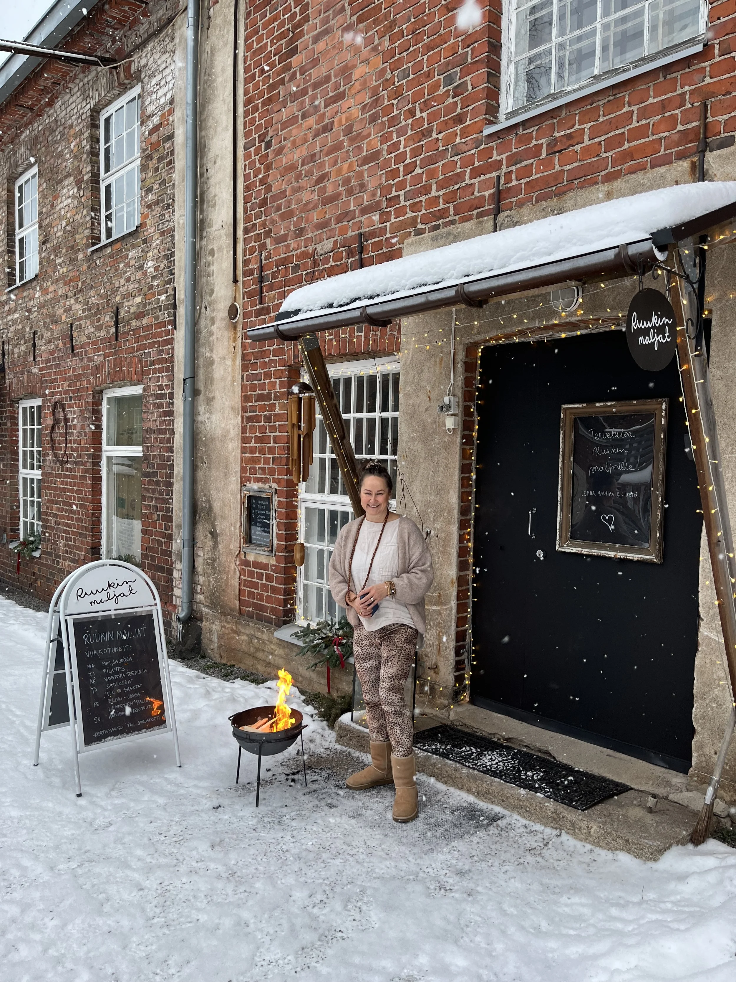 Nainen seisoo lumisella kadulla seinän edessä, jossa on kyltti ja menu taululla. Taasen pieni kaasupoltin sytytettynä ja talvi-asuinen nainen ja rakennuksen tiiliseinät taustalla.