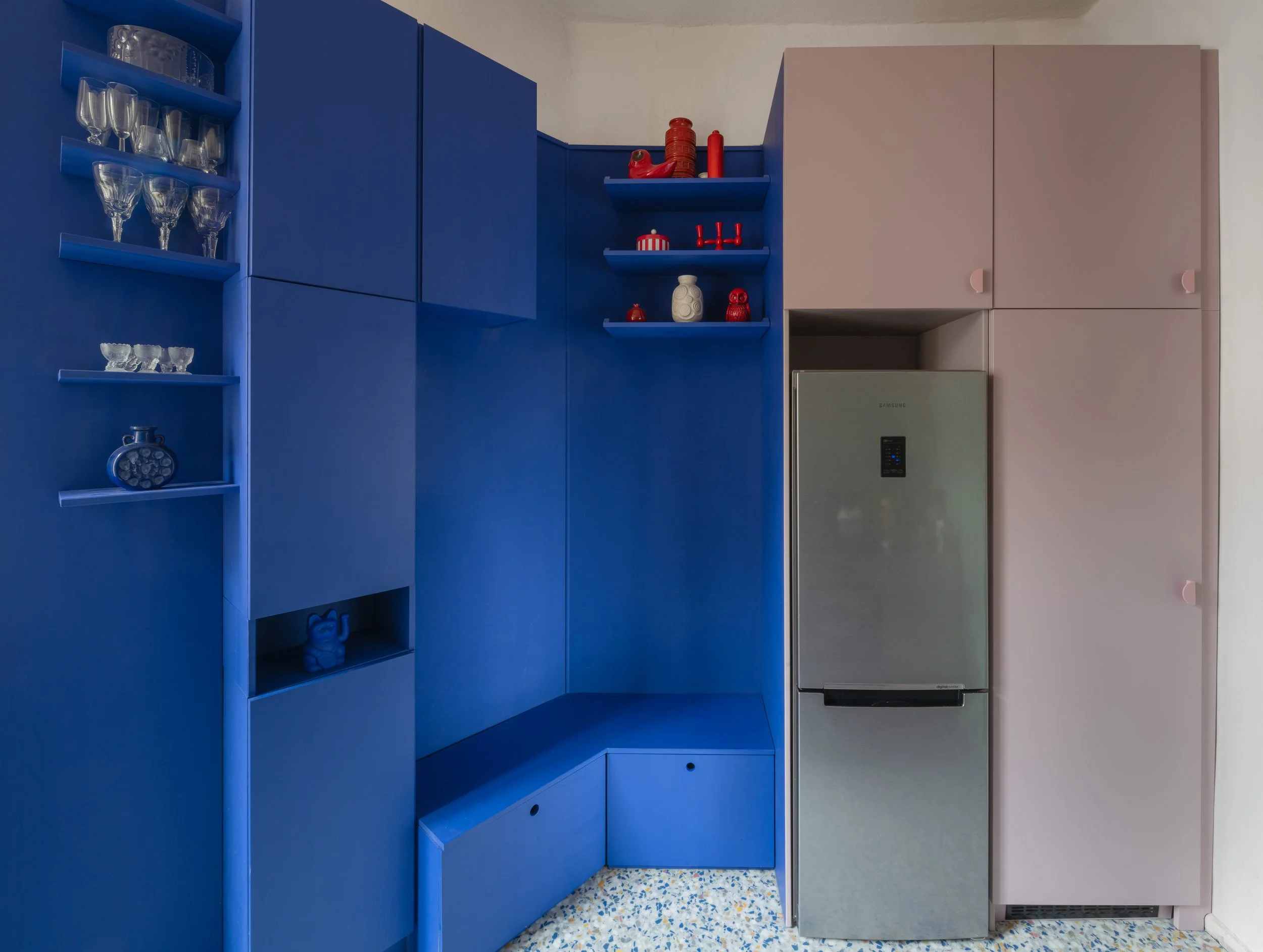 Colorful kitchen cabinet with blue and pink sections, glassware on shelves, a small fridge in an alcove, and decorative items on open shelves.
