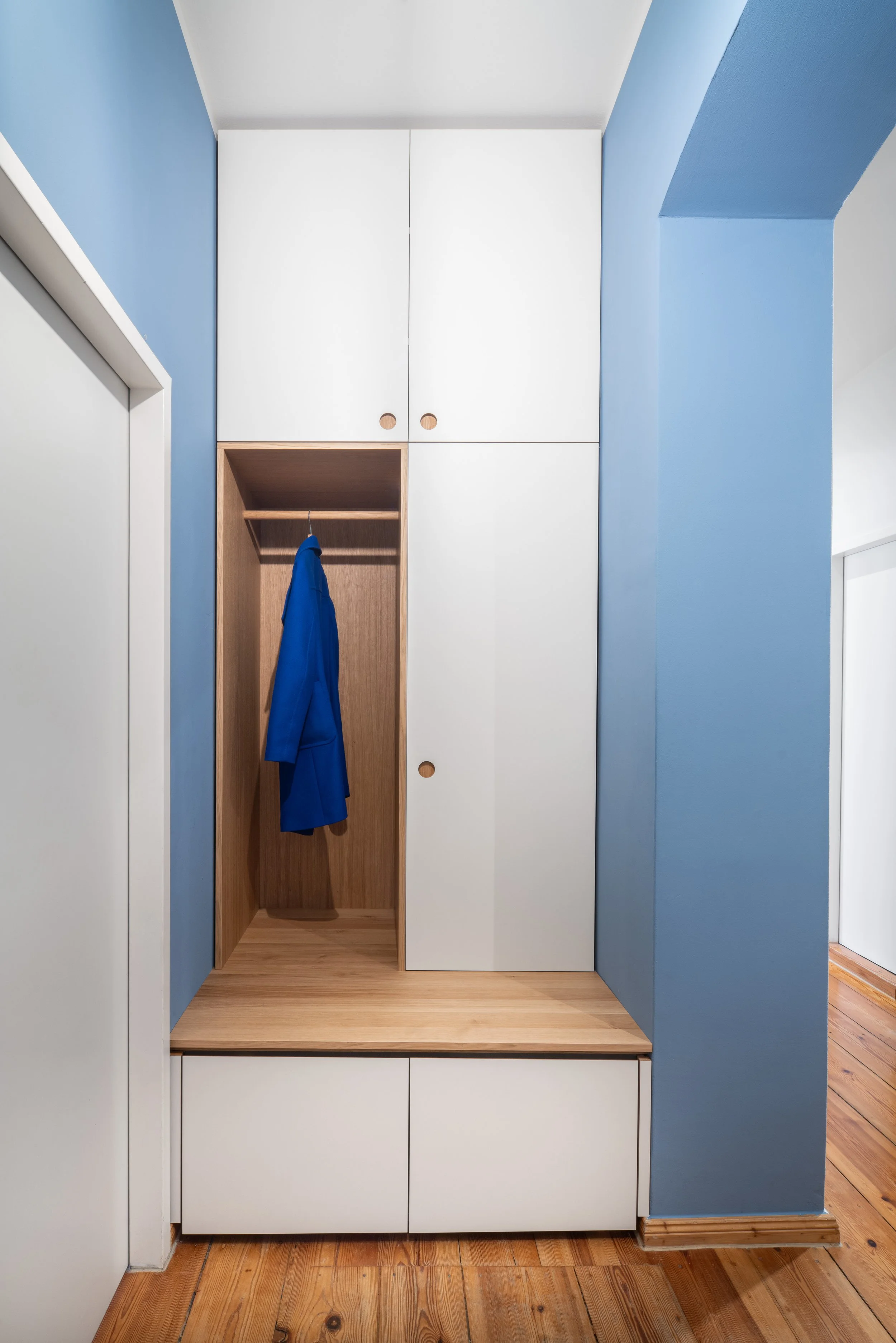 Modern built-in wardrobe with white and wooden doors, blue walls, and a wooden coat rack with a blue coat hanging.