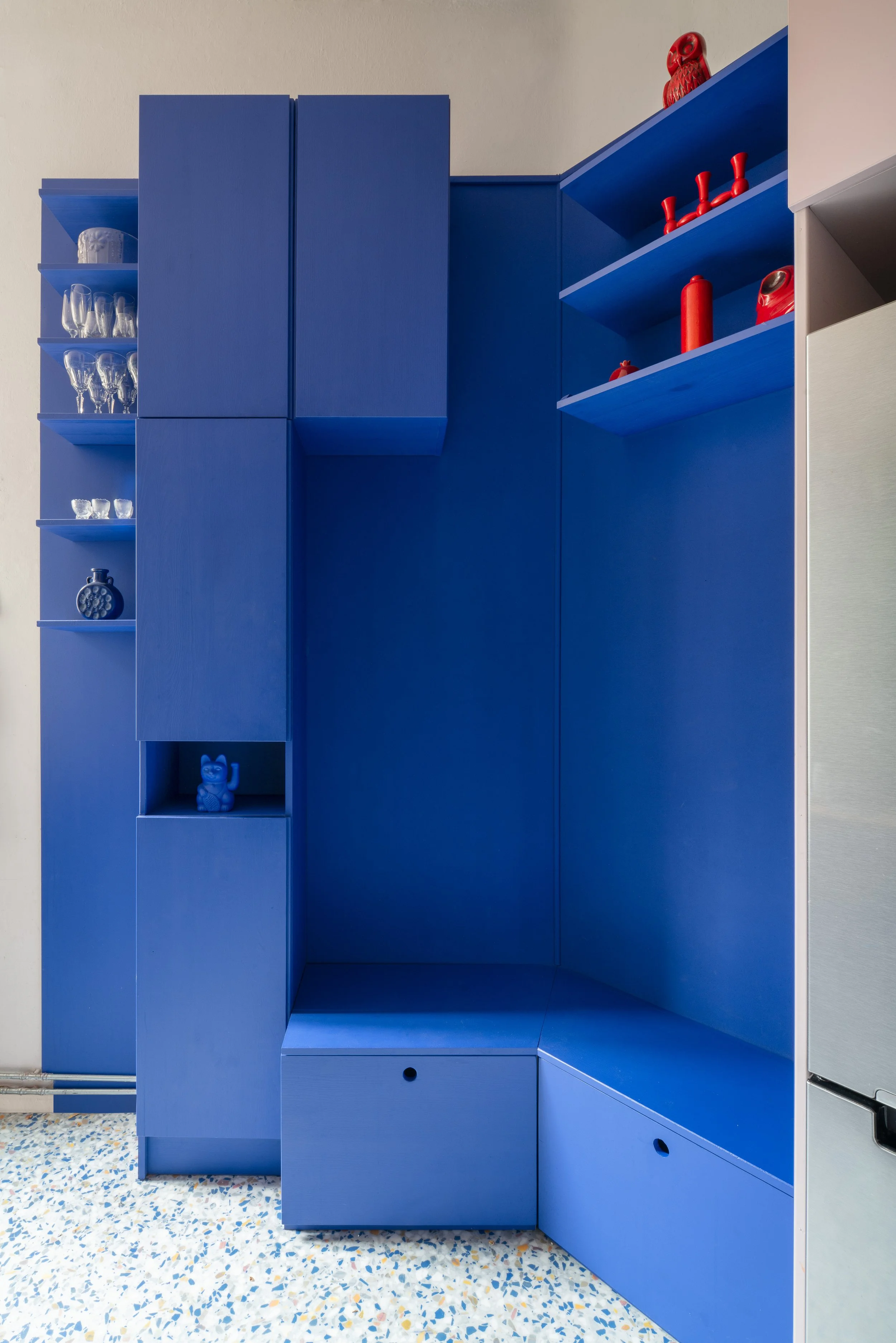 Blue kitchen cabinet with open shelving displaying glassware and decorative items, against a speckled terrazzo floor.