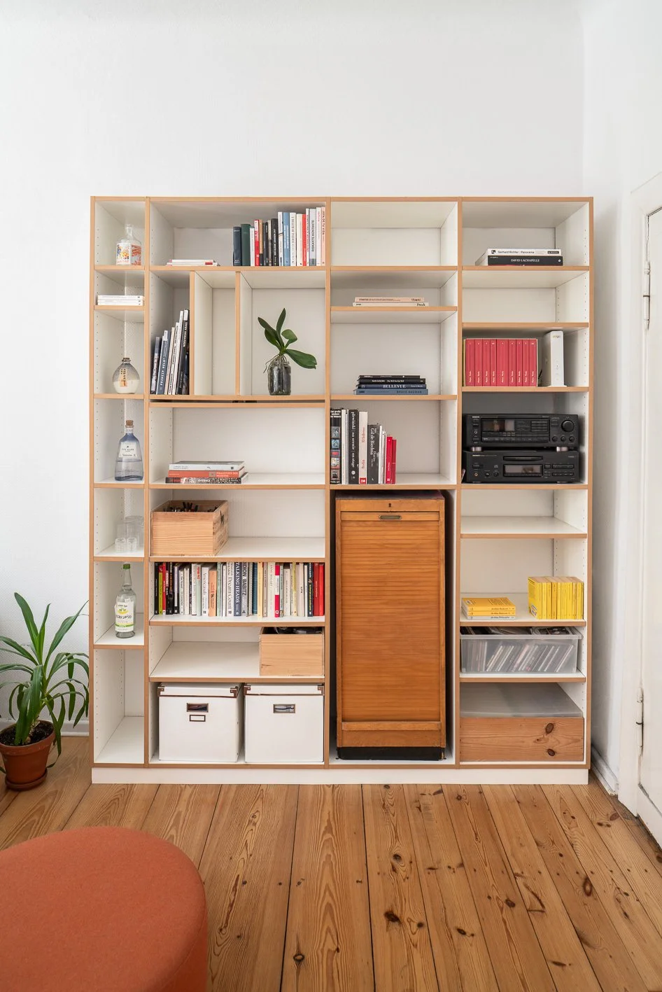 A large white wooden bookshelf with various books, decorative items, a potted plant, a vintage wooden cabinet, and storage boxes, set on a wooden floor next to a white wall and a door.
