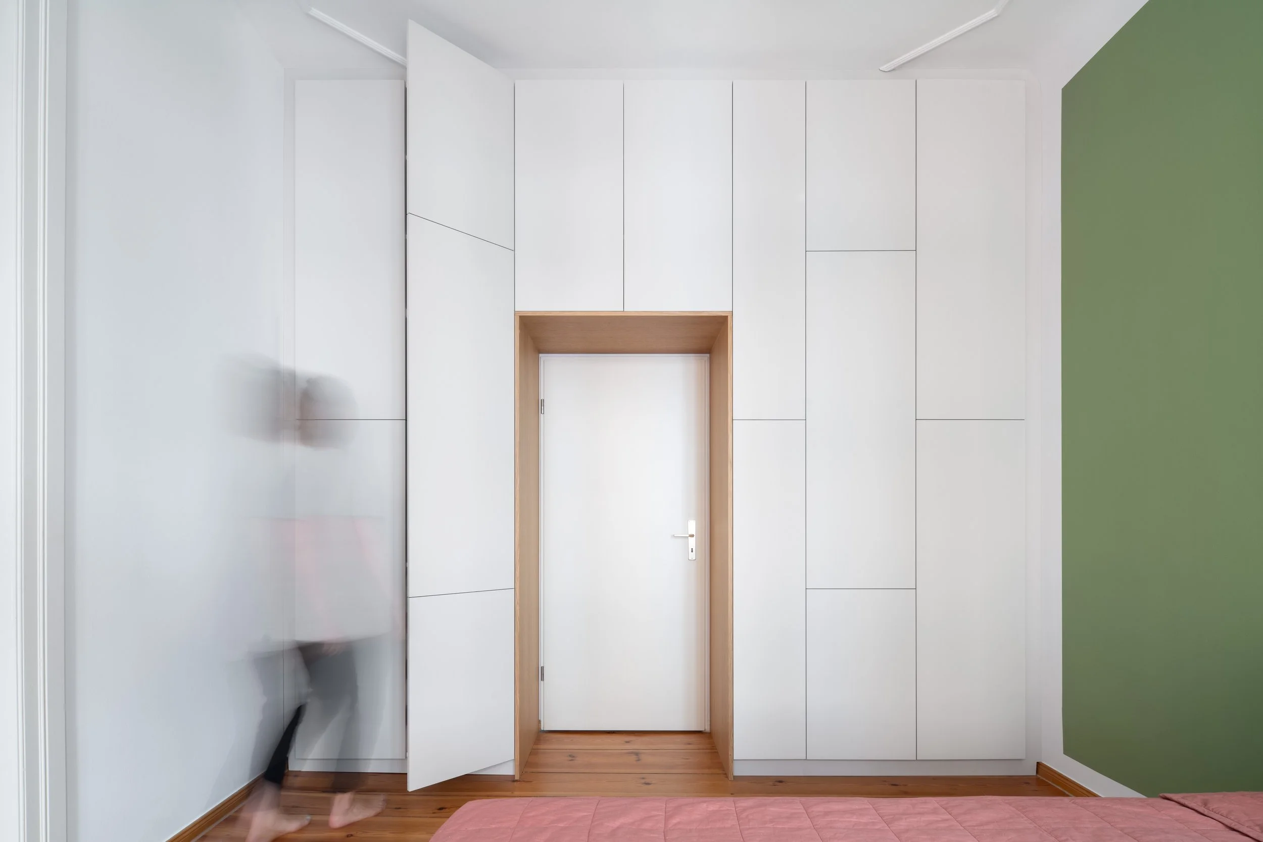 A bedroom with a white wardrobe, a white door with a wooden frame, and a green accent wall. The floor is wood, and a pink bedspread is partially visible at the bottom of the image.
