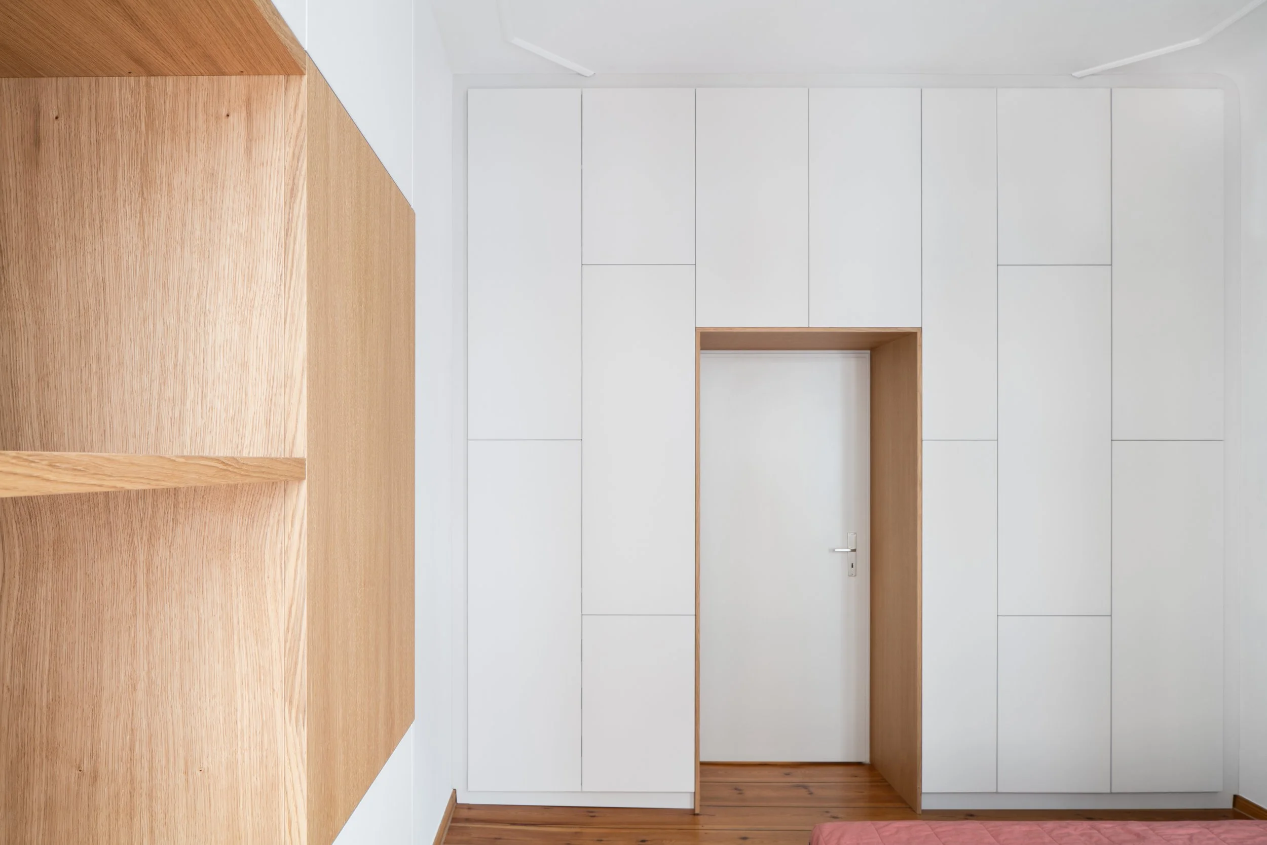 Interior view of a modern room with a white wall, wooden flooring, a white door framed in wood, and a wooden shelf on the left side.