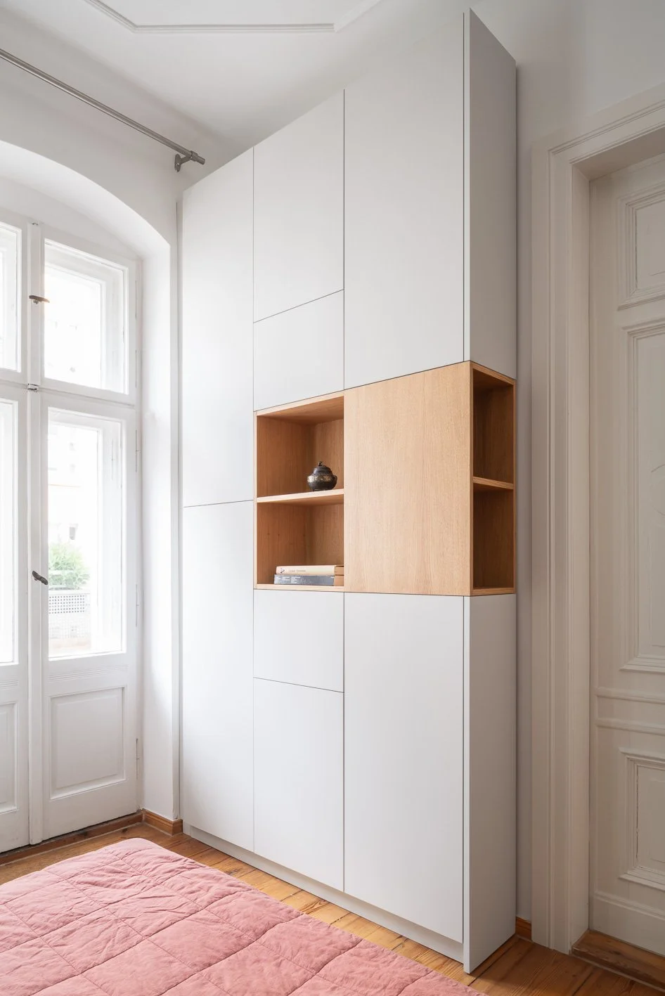 A tall, white modern wardrobe with wooden open shelves in a room with a window and a white door.