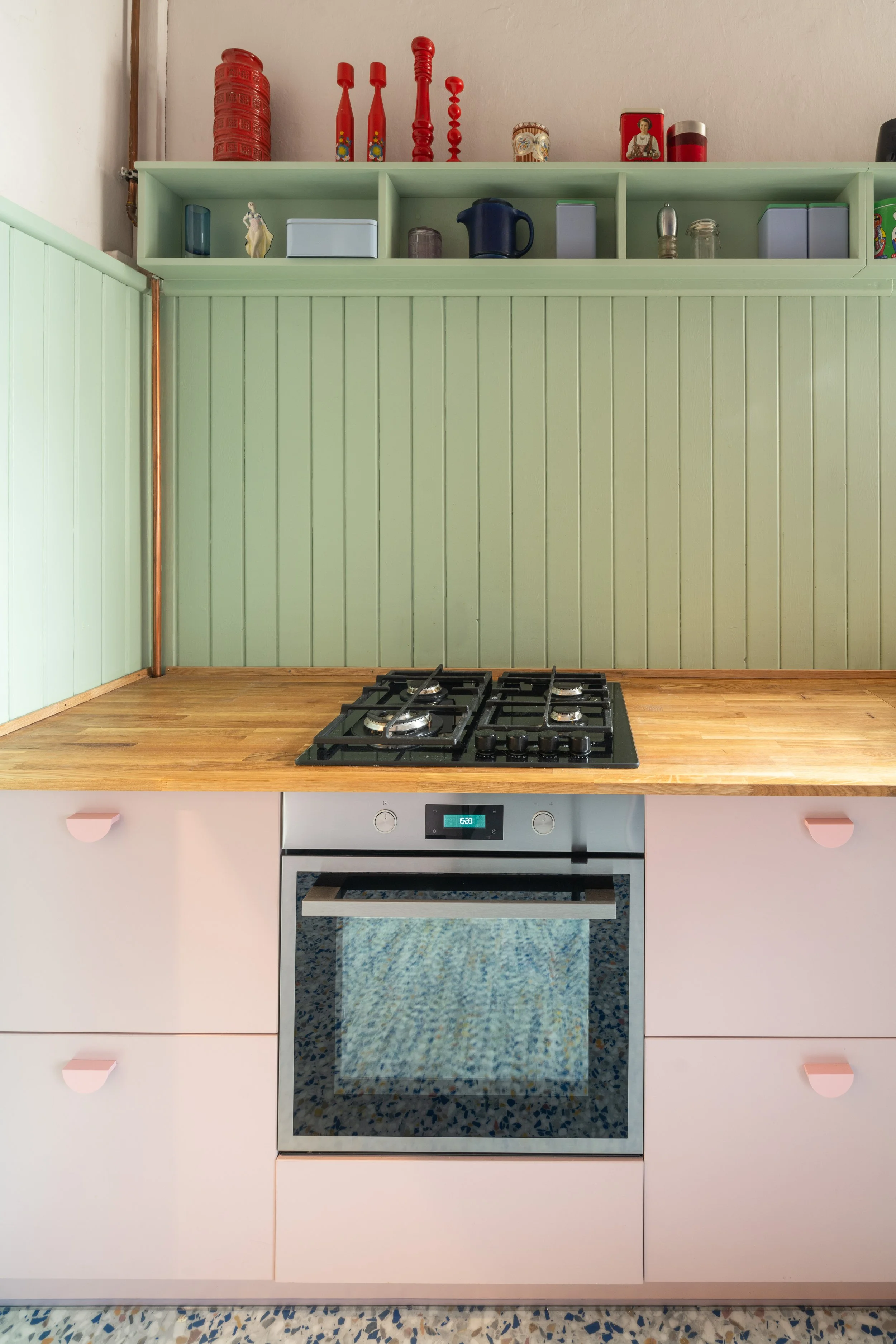 A kitchen with pastel pink cabinets, a wooden countertop, a built-in oven, and mint green wall paneling with open shelves holding various jars, containers, and decorative items.