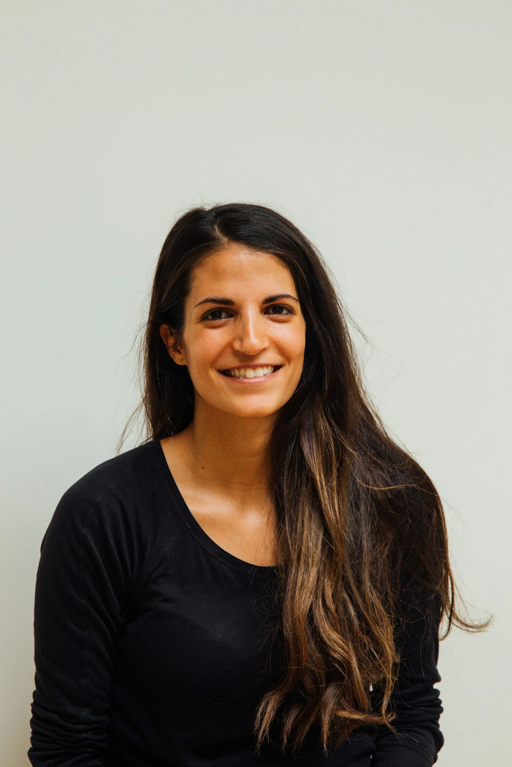A young woman with long, dark hair, smiling, wearing a black long-sleeve shirt standing against a plain light-colored wall.