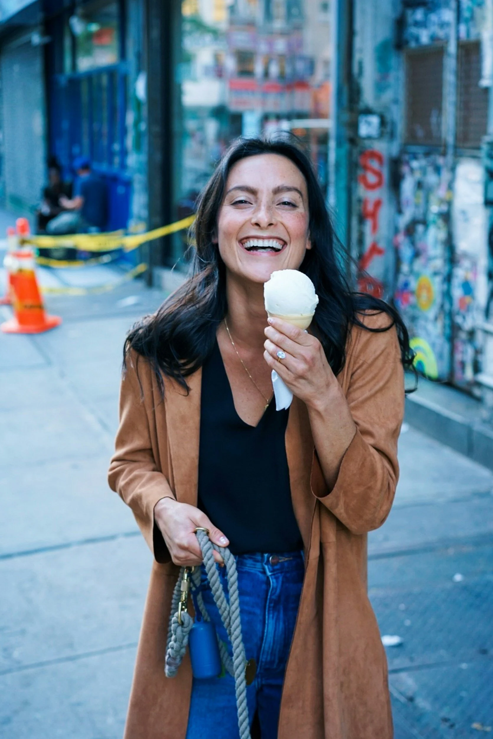 A woman smiling and holding a single scoop of vanilla ice cream in a cone, standing on a city sidewalk with graffiti-covered walls and a few people in the background.