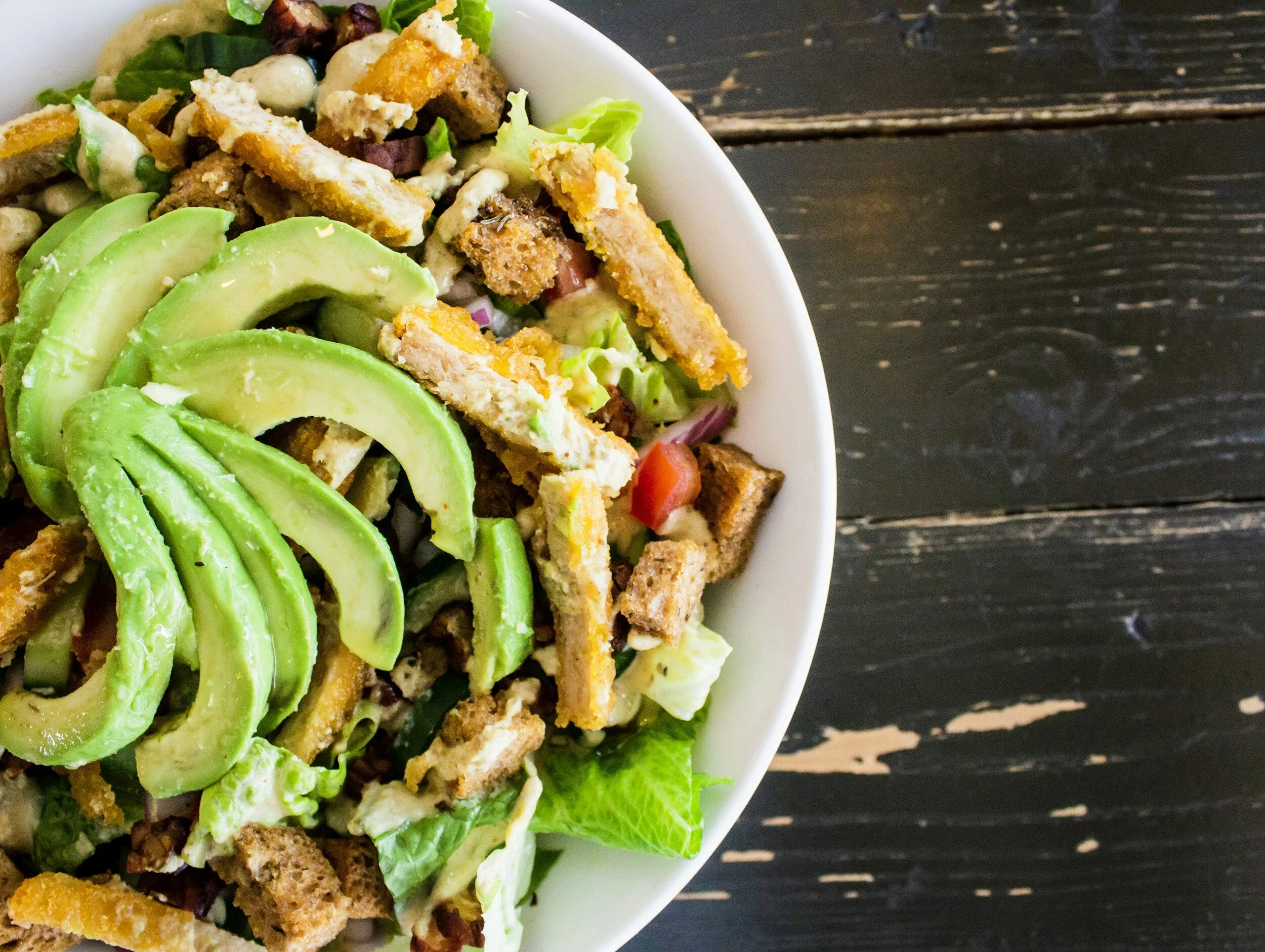 A white bowl containing a salad with sliced avocado, crispy fried tofu, chopped lettuce, and other vegetables on a dark wooden surface.