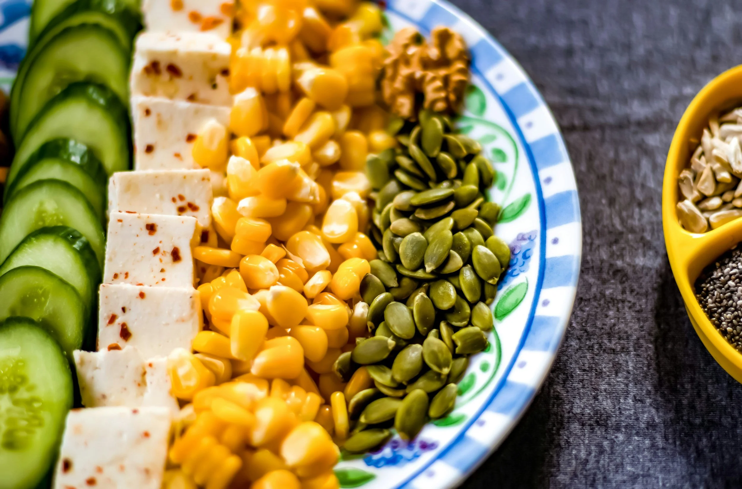 Plate with sliced cucumbers, cubed tofu with red pepper flakes, corn, and pumpkin seeds on a decorative blue and green plate.