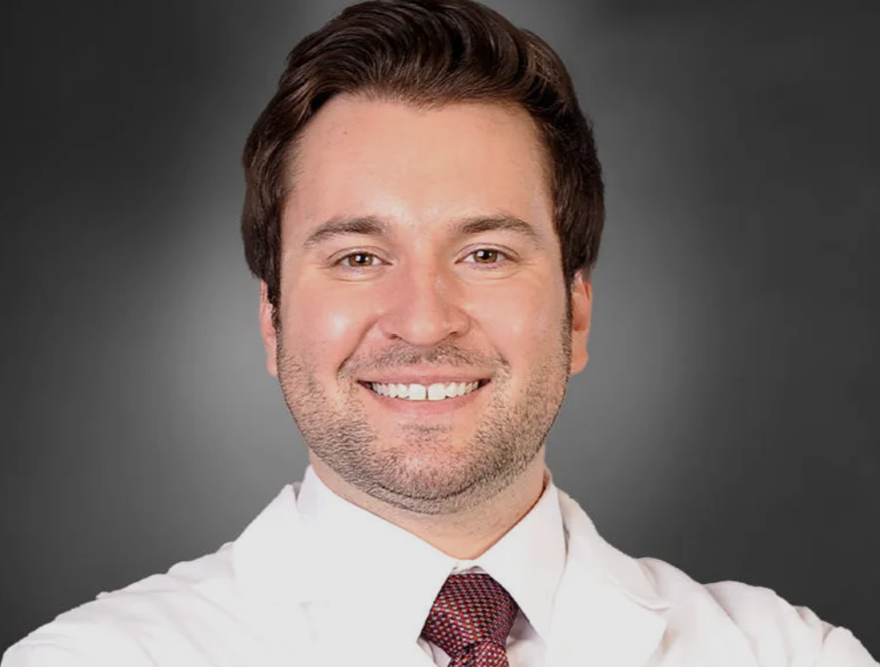 A smiling man with dark brown hair, wearing a white coat and a red tie, against a gray background.