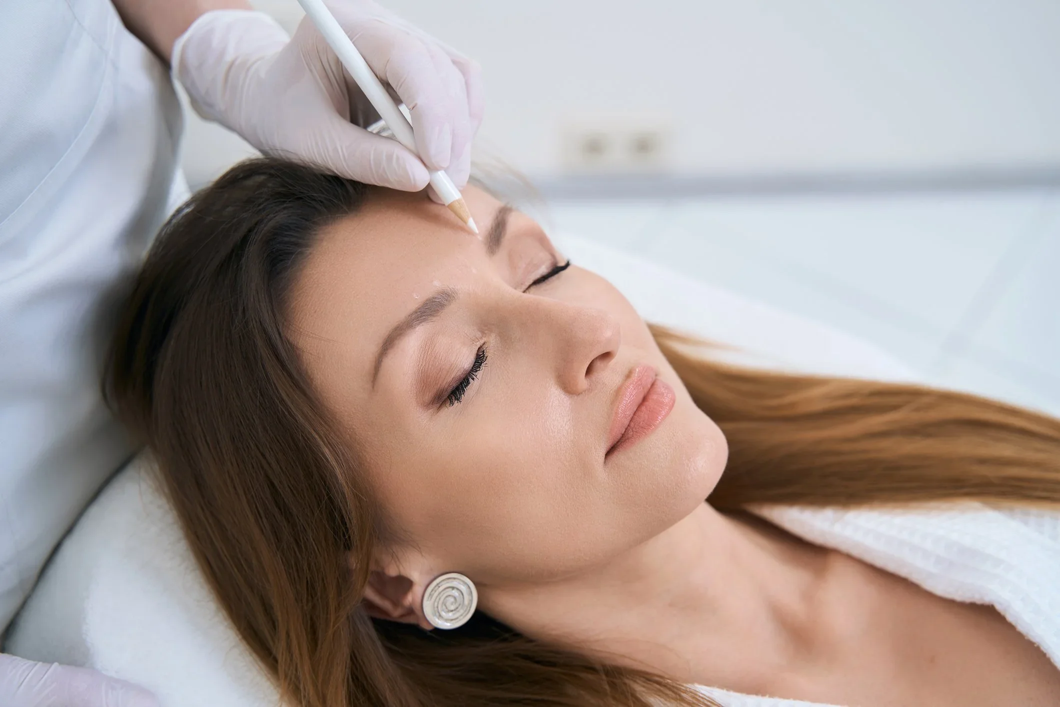 A woman with closed eyes lying on a treatment bed while a person in gloves uses a pinpoint tool to treat her forehead.