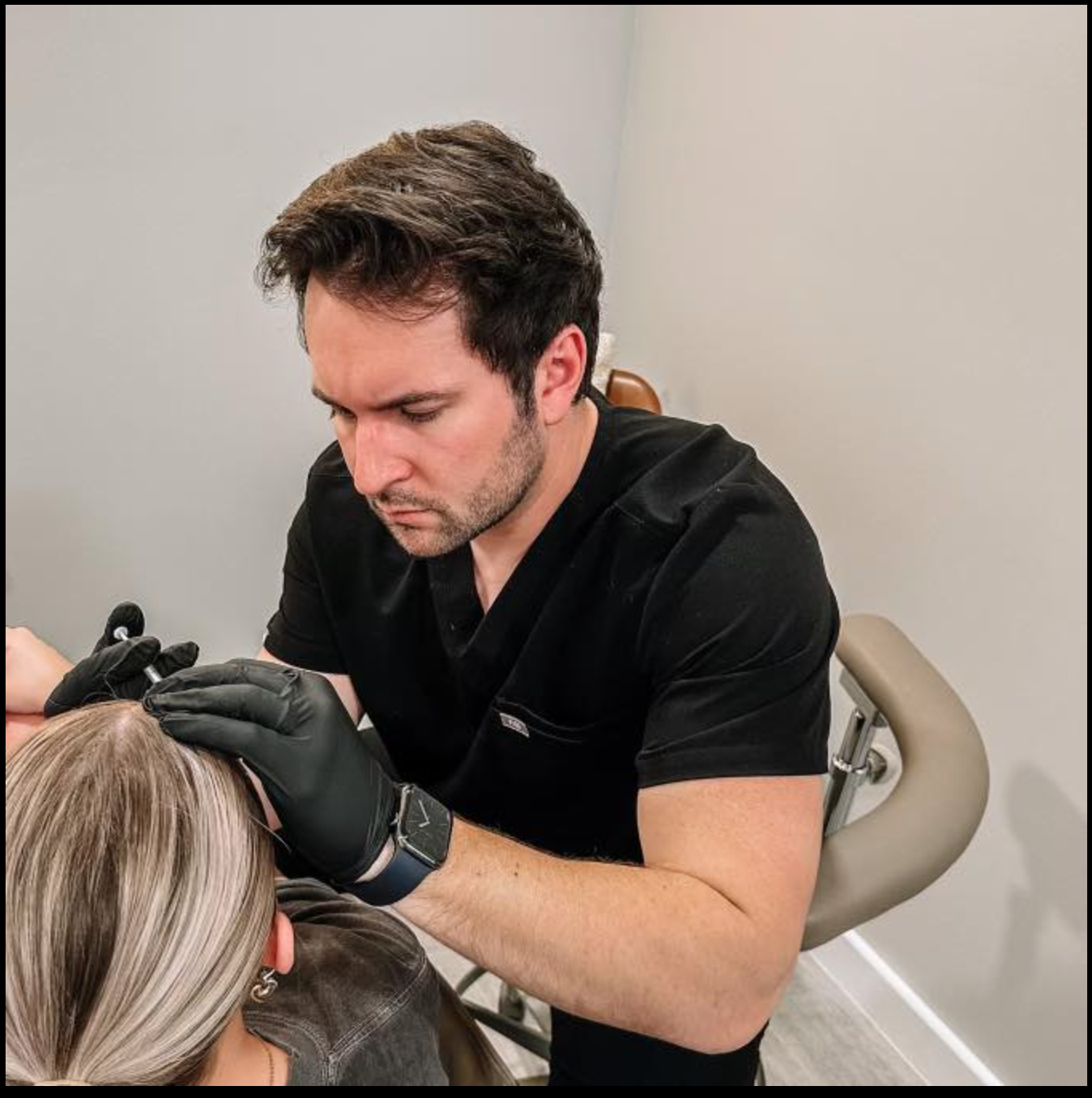 A tattoo artist with dark hair, wearing black scrubs and black gloves, is focused on creating a tattoo on a woman's arm in a tattoo studio.