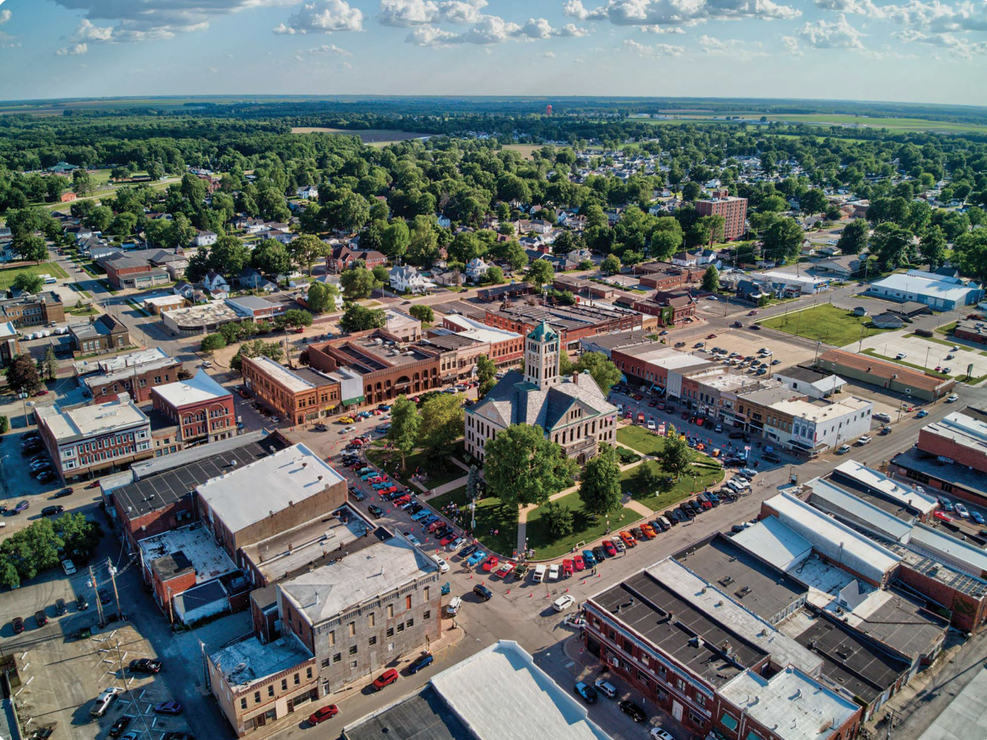 Aerial view of a small town with a central courthouse, surrounded by streets, shops, and houses, with a mix of trees and buildings extending into the horizon under a partly cloudy sky.