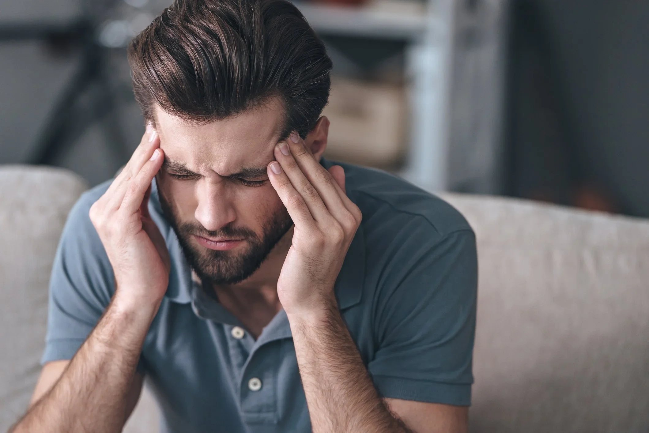 A man with dark hair and a beard holding his head with both hands, looking distressed or in pain, sitting on a couch.
