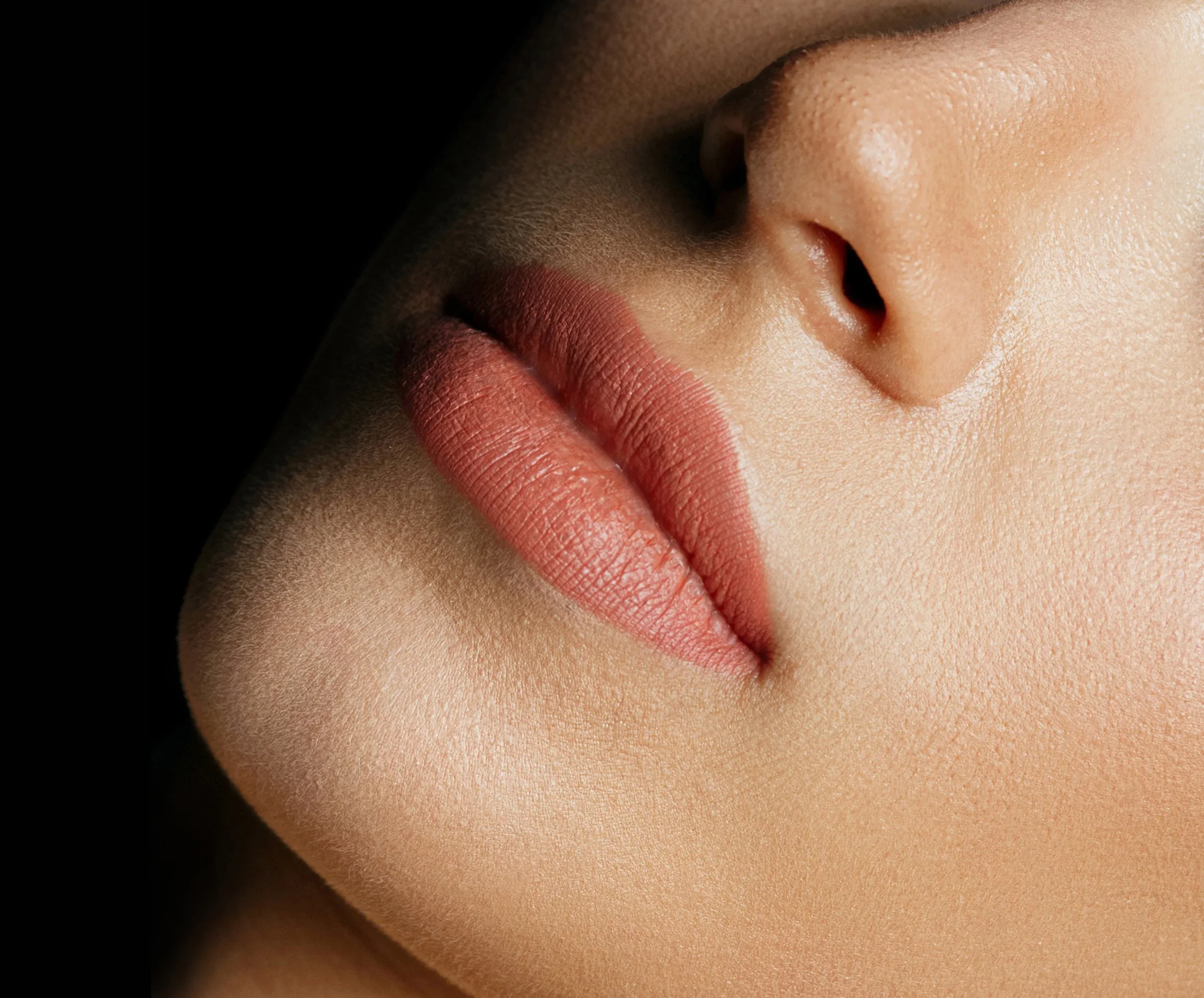 Close-up of a woman's lips with matte lipstick, her nose, and part of her cheek, with smooth skin and soft lighting.
