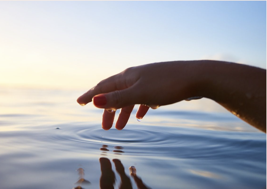 A hand with red-painted nails touching water at sunset, creating ripples.
