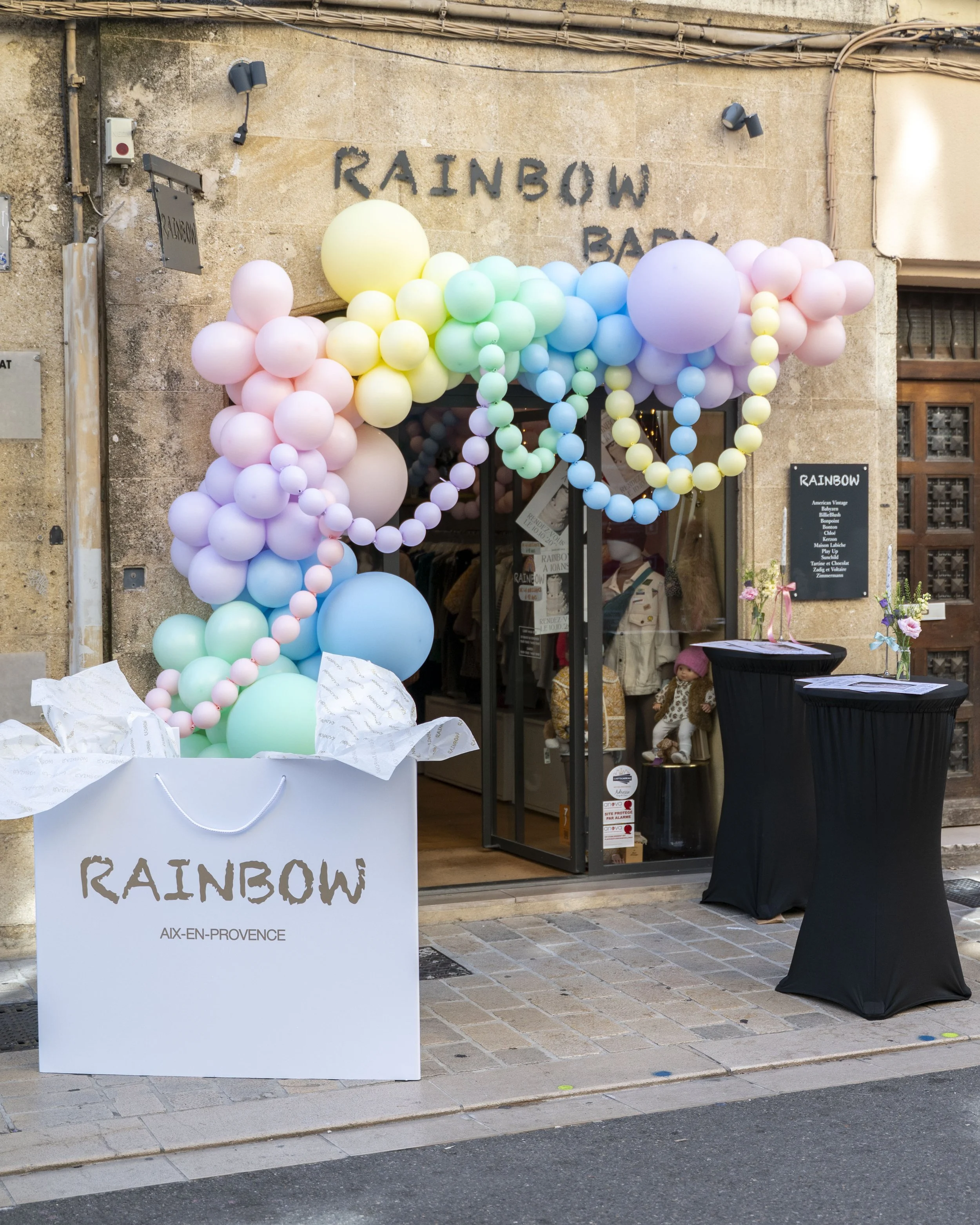 Vitrine décorée avec des ballons de différentes couleurs formant une arche, devant une boutique appelée Rainbow à Aix-en-Provence, avec une grande boîte blanche portant le nom Rainbow et deux tables hautes avec des fleurs.