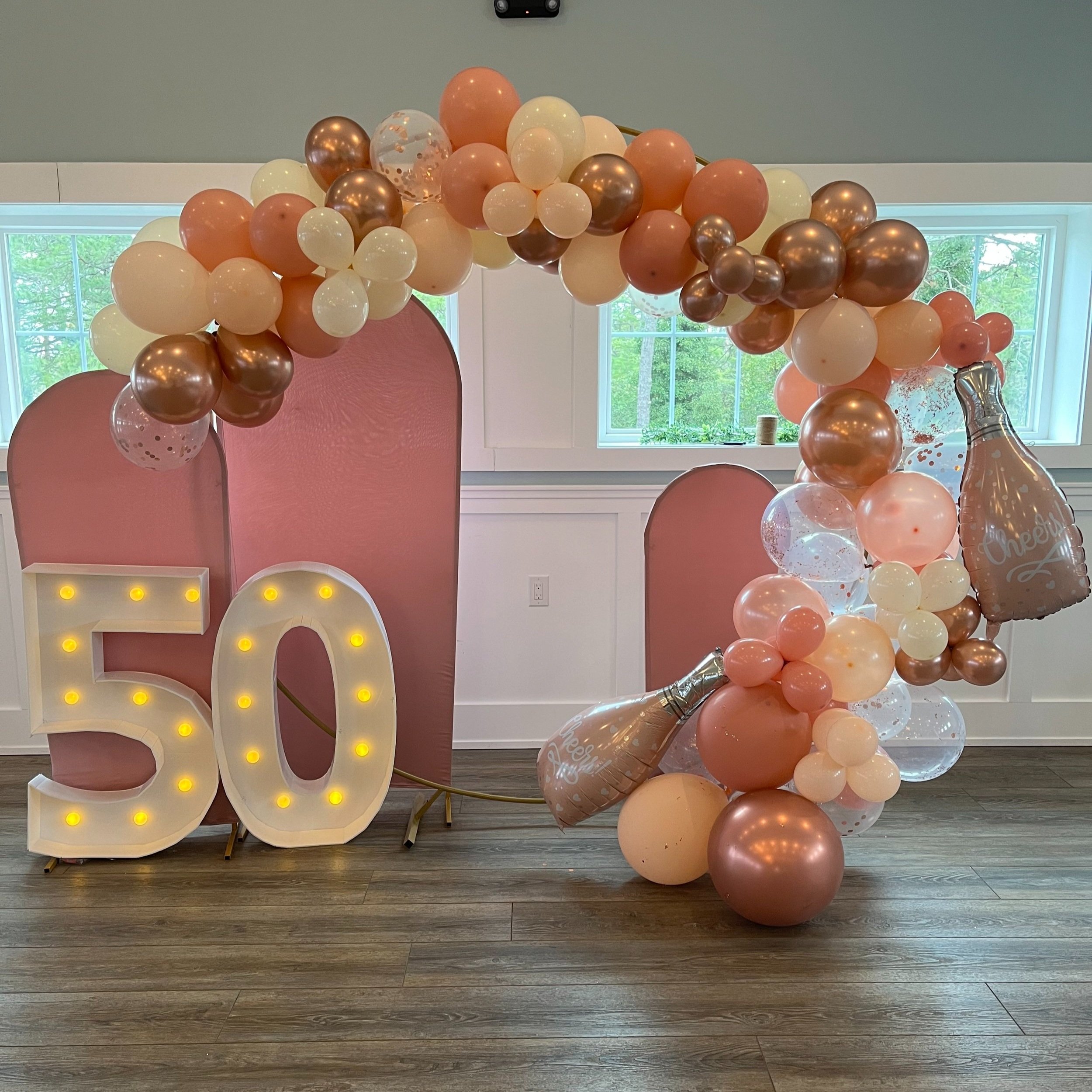 Decorative 50th birthday celebration setup with a pink backdrop, illuminated '50' sign, and an arch of pink, white, and rose gold balloons, including champagne bottle-shaped balloons with 'Cheers' written on them.