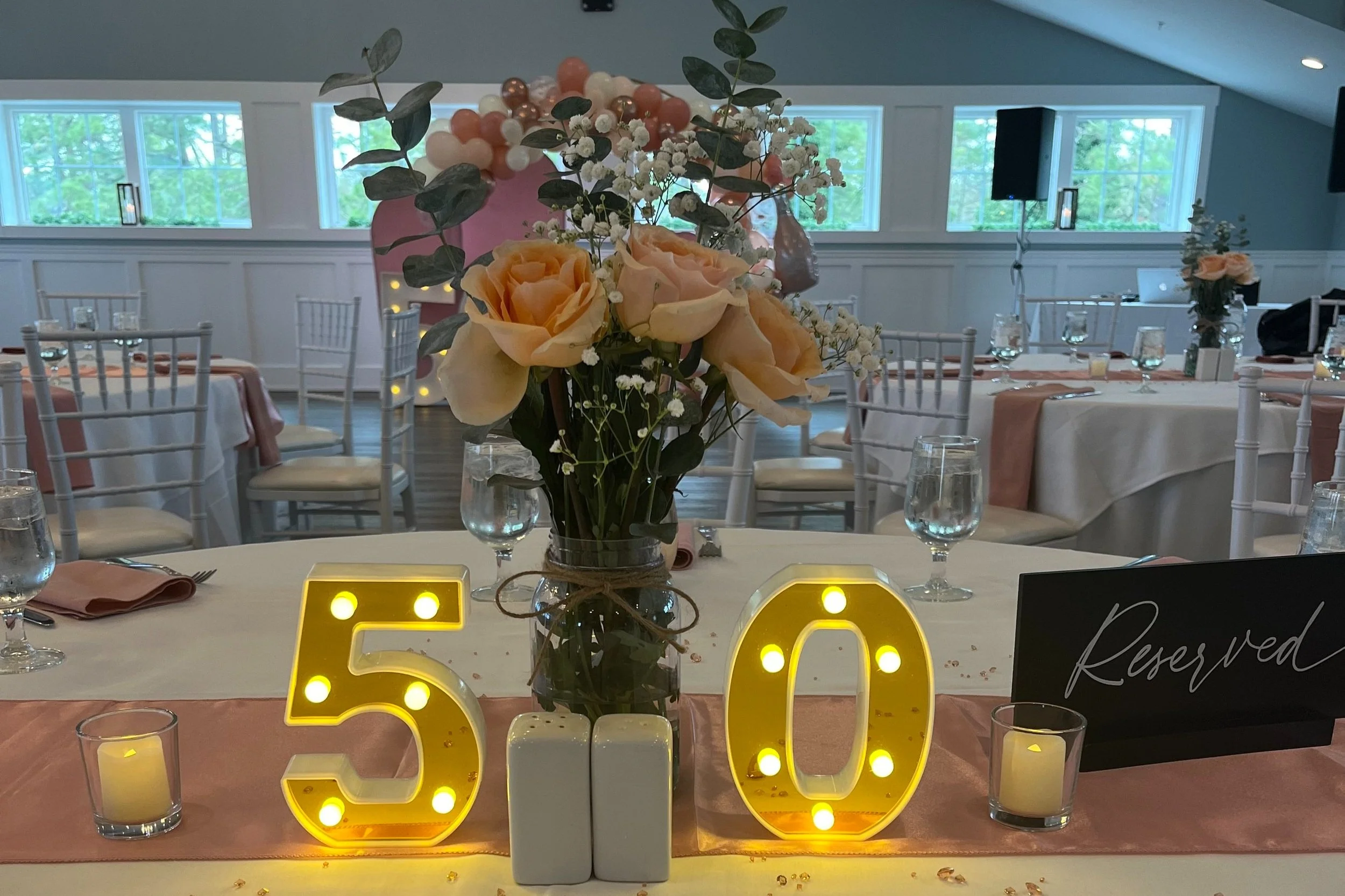 Wedding reception table decorated with a floral centerpiece, illuminated '50' table number, reserved sign, candles, and table settings with pink and white accents.
