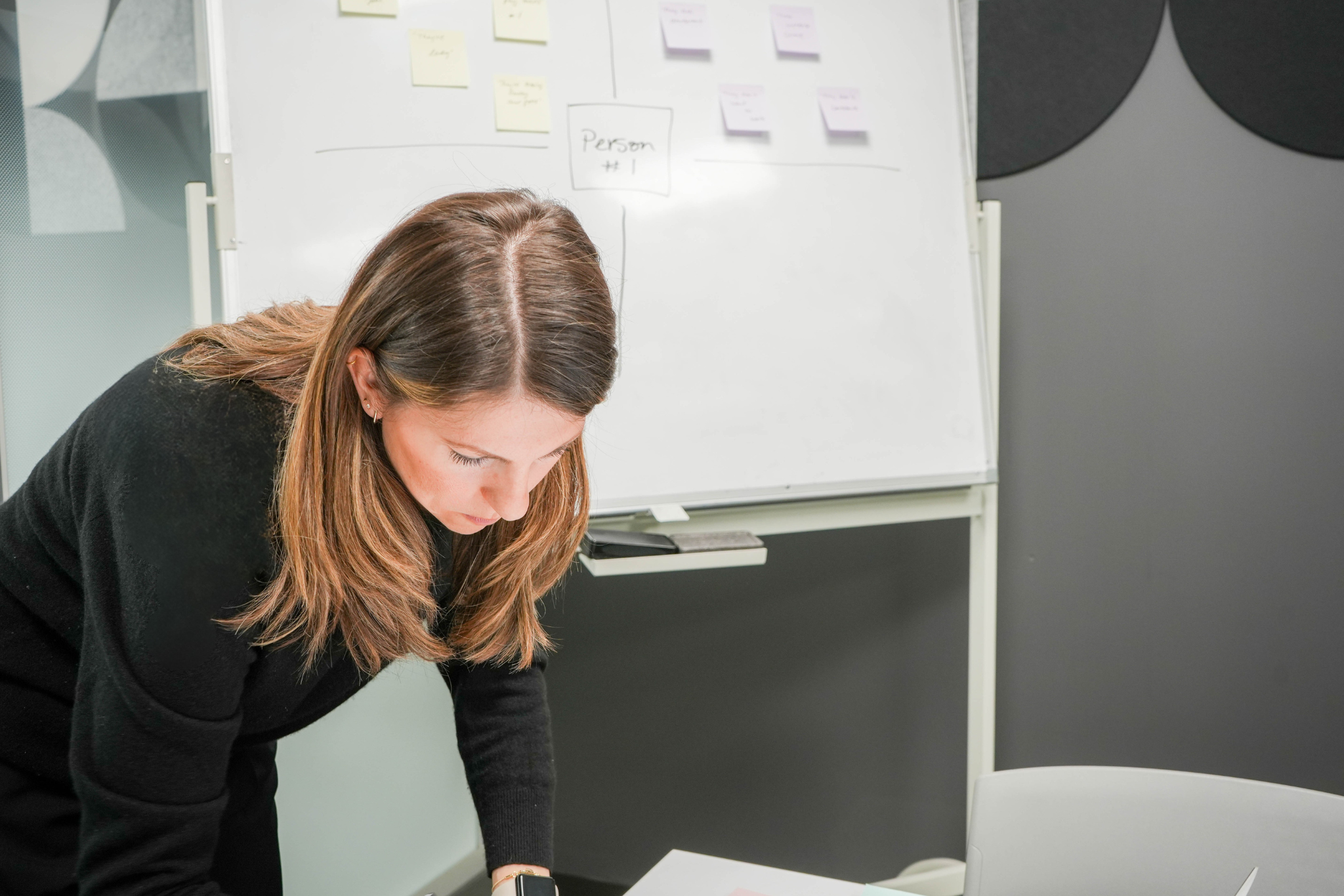 A woman with brown hair leaning over a table looking down at something off-screen. In the background, there is a whiteboard with Post-it notes and sketches.