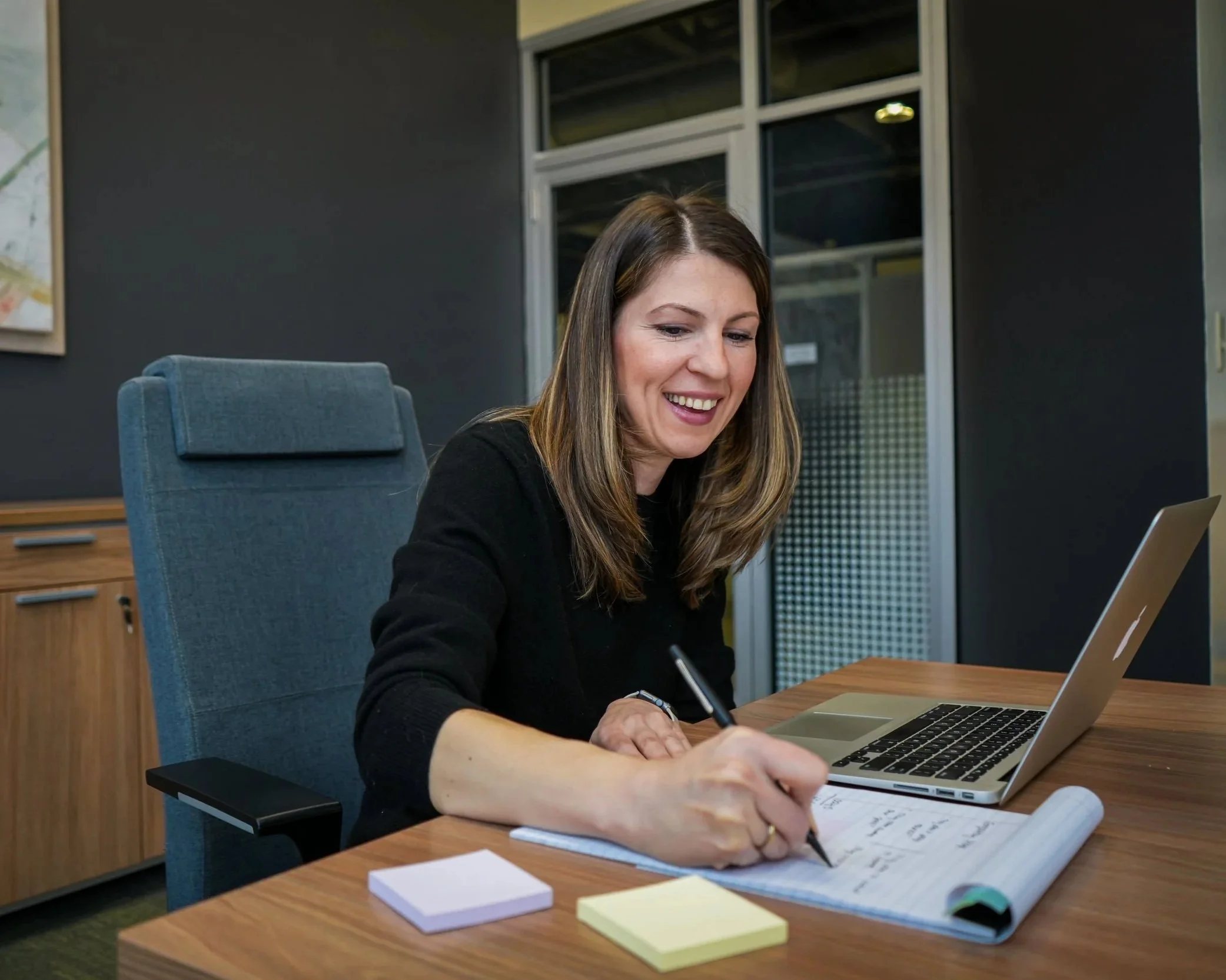 A woman sitting at a desk, smiling, writing notes, with a laptop open in front of her.