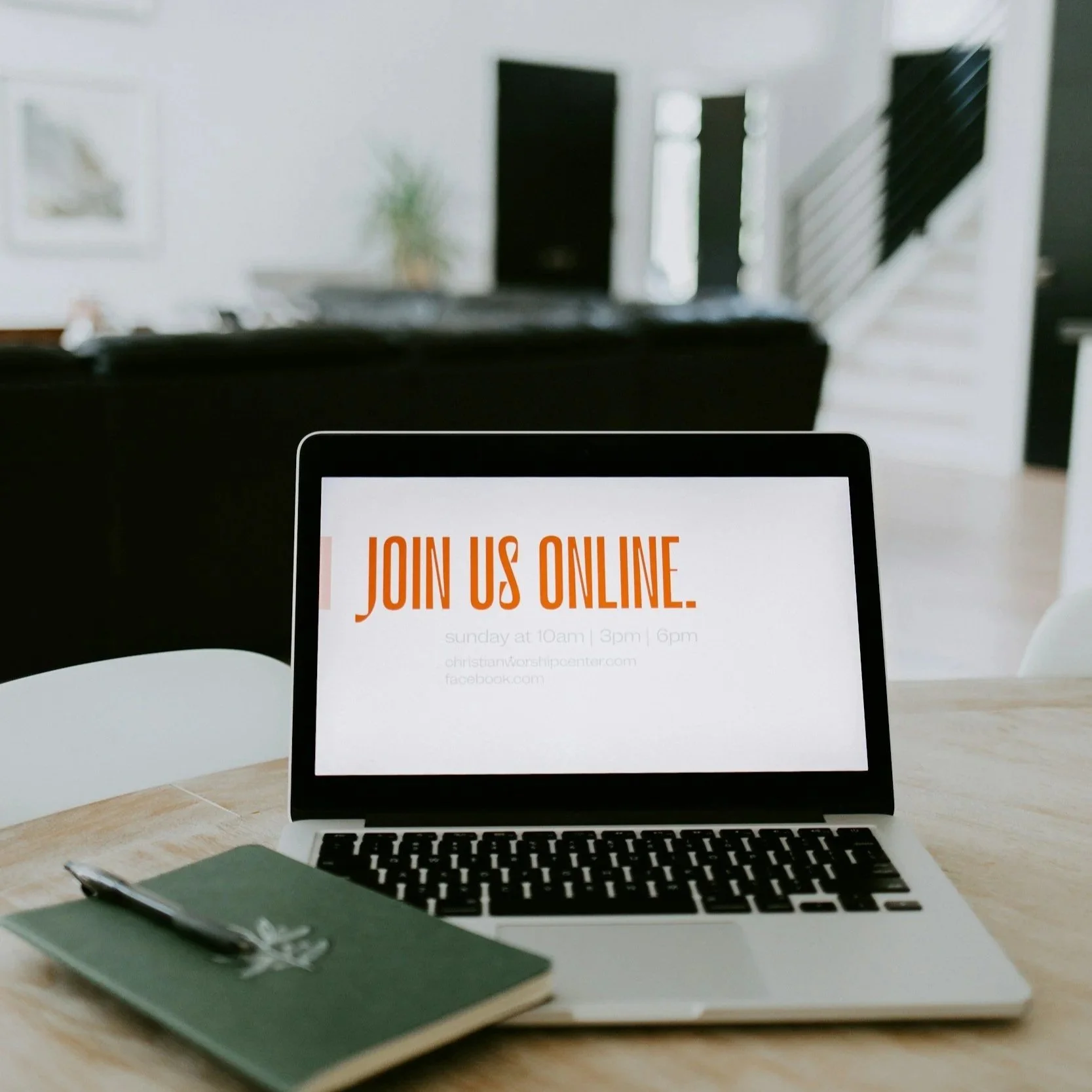 Laptop on a wooden table displaying a sign that says 'Join us online,' with additional details about times and website. A closed green notebook and pen are placed next to the laptop.