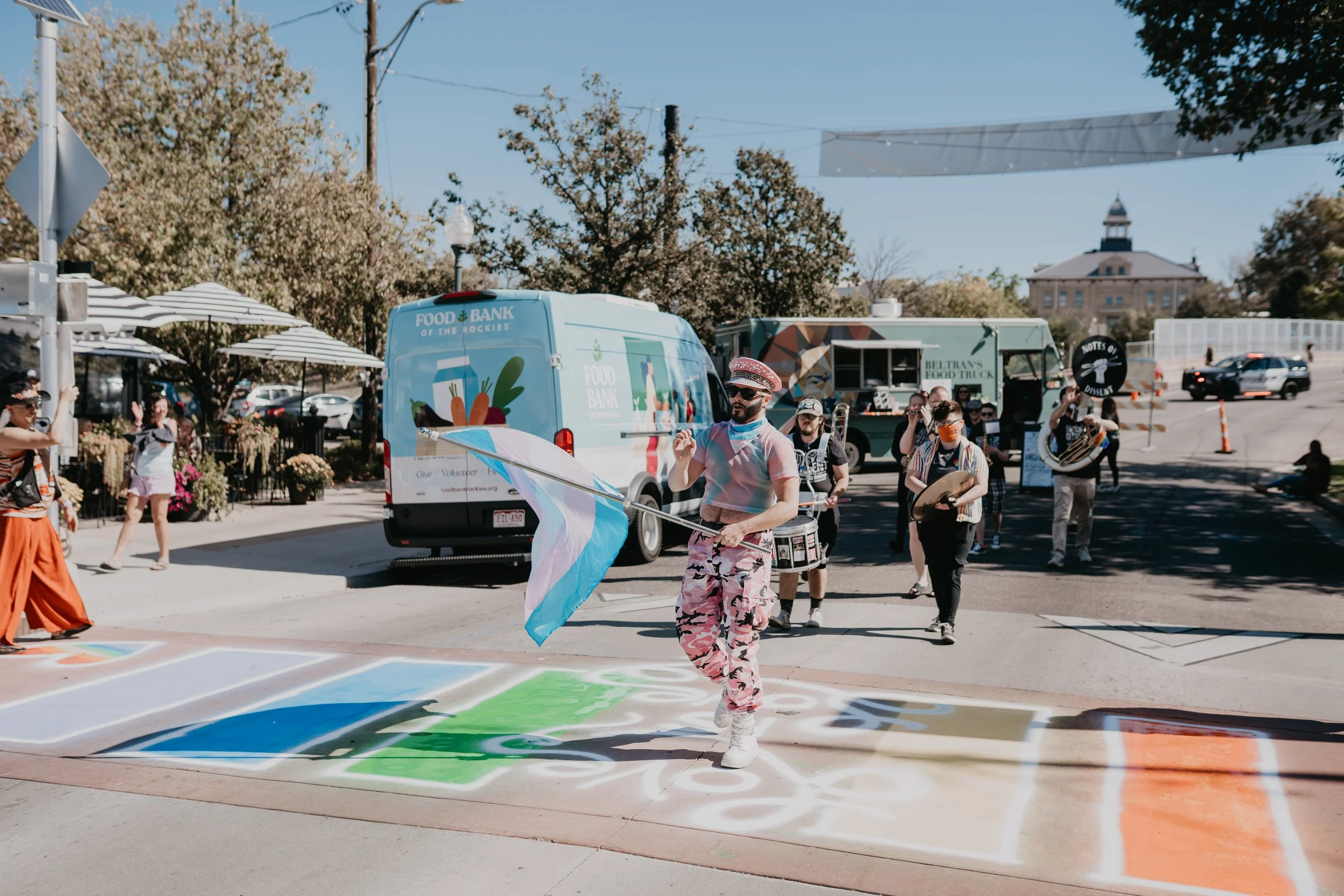 Colorful street parade with musicians, dancers, food trucks, and a person carrying a transgender pride flag.