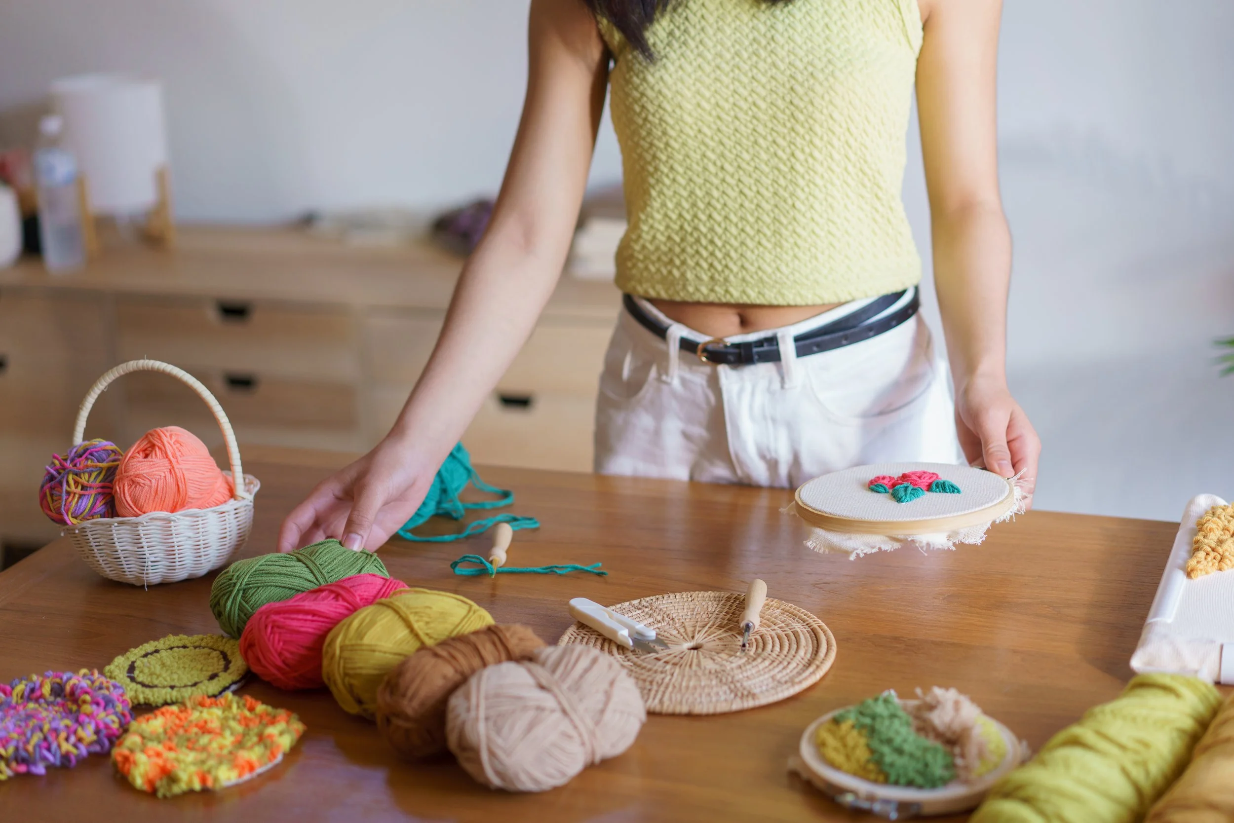 A person in a yellow knit sweater standing at a table with multiple colors of yarn balls, knitting tools and an embroidery hoop