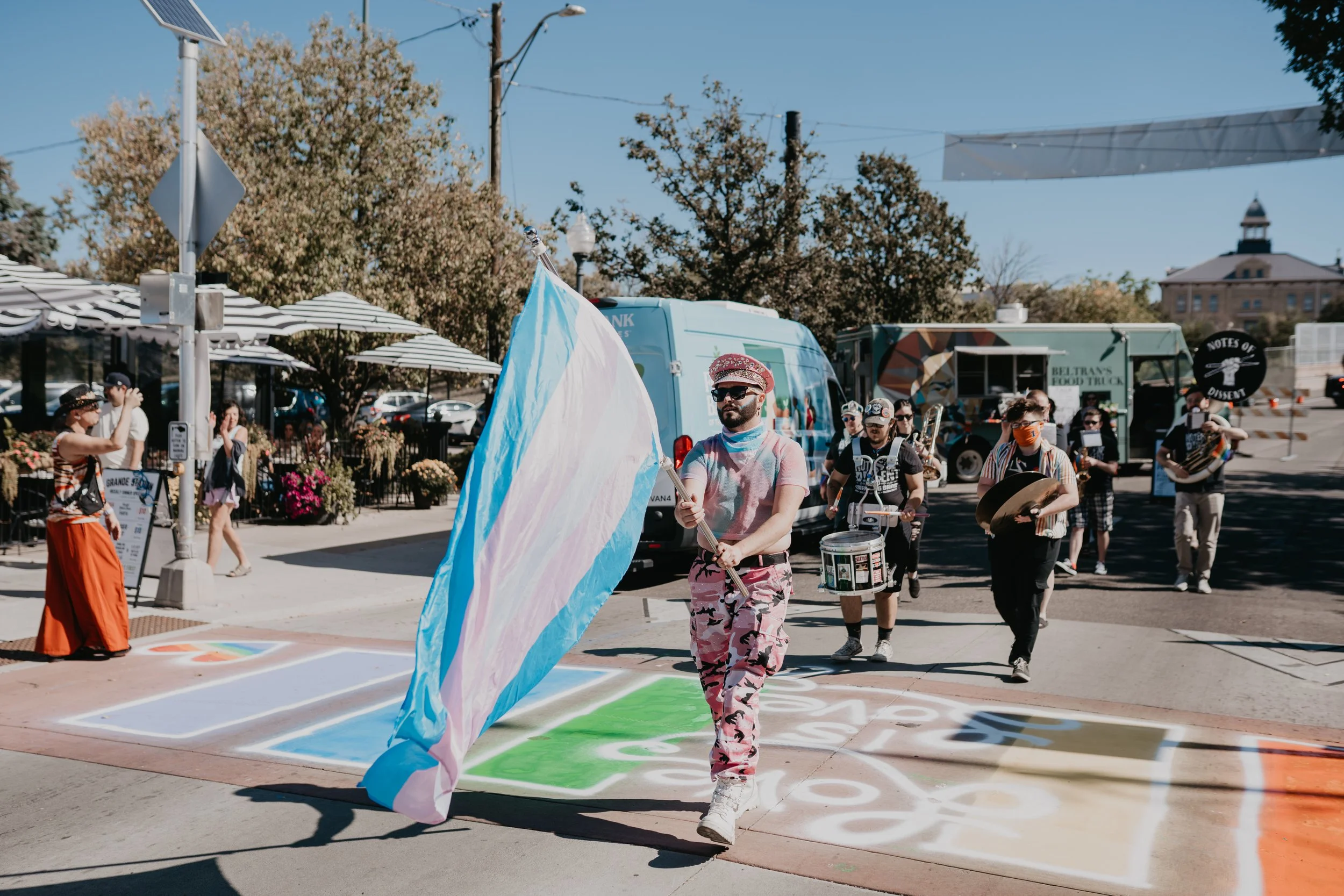 Pride parade participants marching with a transgender pride flag and drums on a colorful downtown street