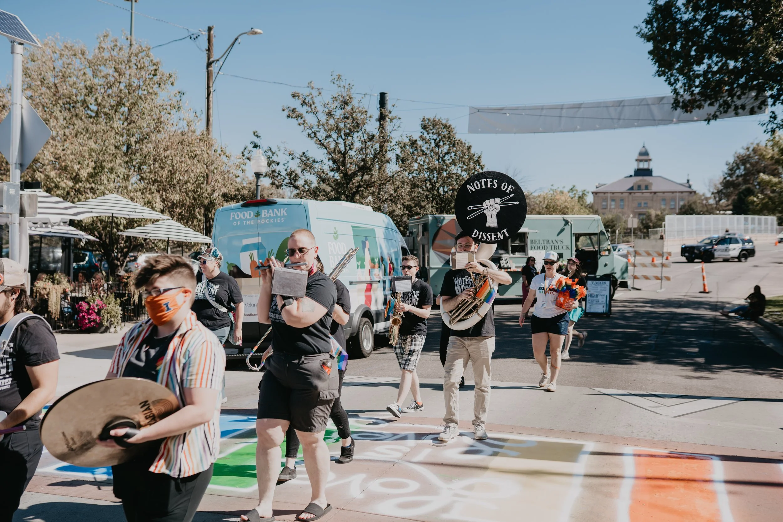 Masked musicians march in a colorful street parade past a Food Bank of the Rockies van and food trucks.