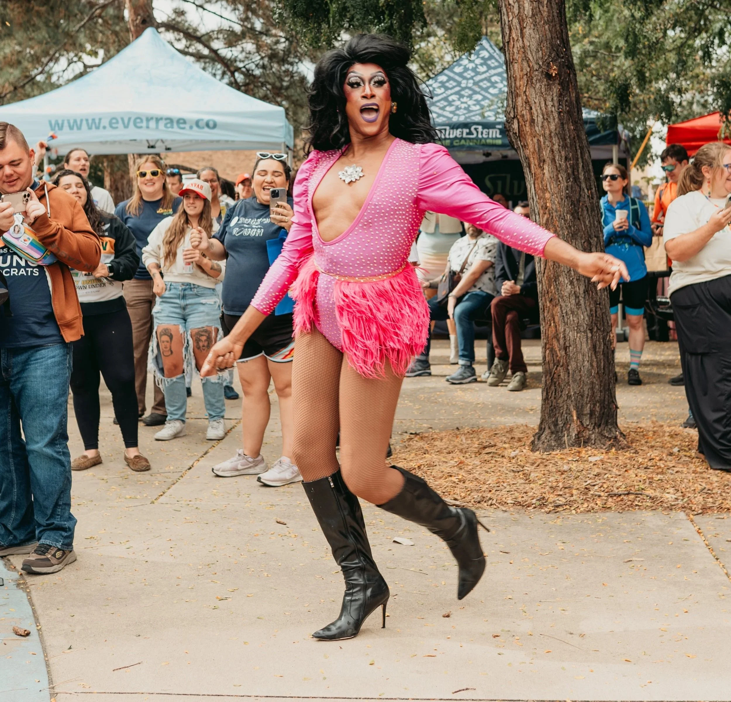 Drag Queen, Juiccy Misdemeanor, in a pink dress and knee high boots dances in the park surrounded by people.