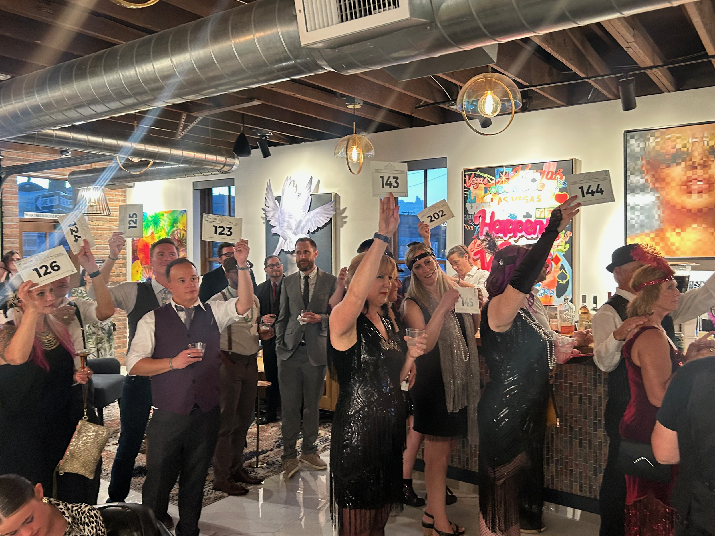 People dressed in 1920s-style costumes with headbands and fringe, holding auction paddle cards with numbers, at a themed party in a decorated indoor venue.