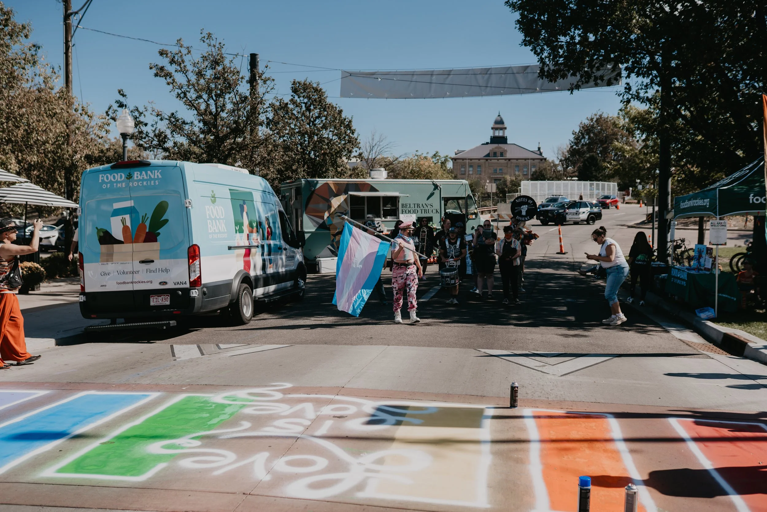 Outdoor community event with food trucks, a Food Bank of the Rockies van, Pride flags, colorful sidewalk art, and a historic building.