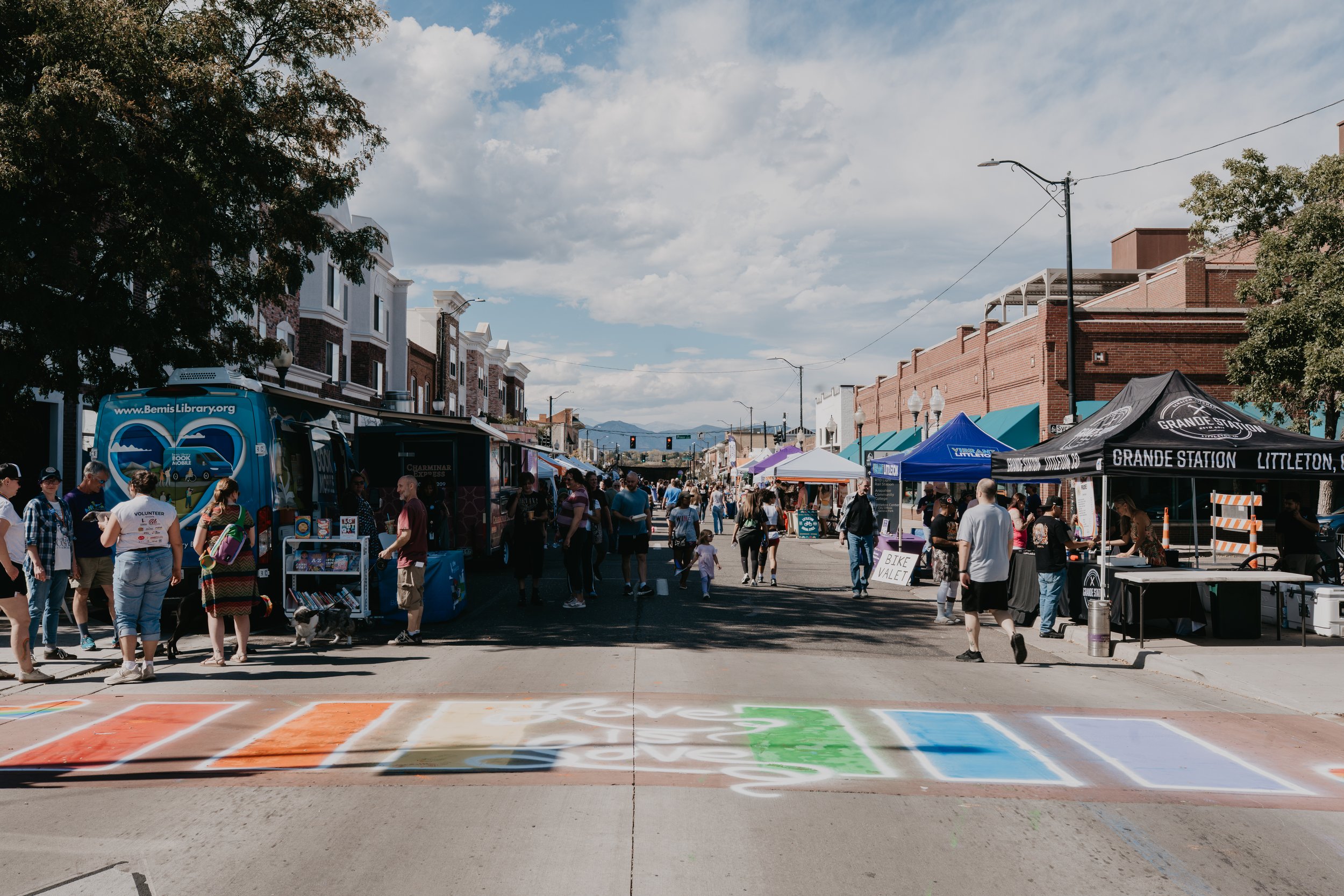Crowded street festival in downtown Littleton, Colorado, with vendor tents, a Bemis Public Library bookmobile, colorful street art, and mountains in the background.