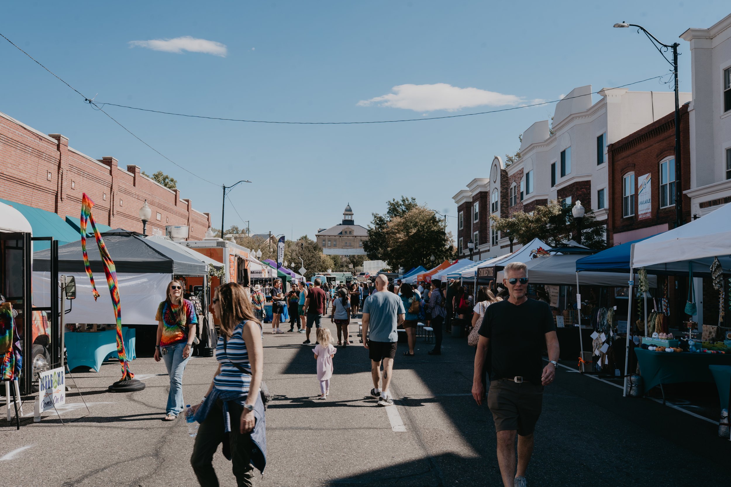 Crowded downtown street festival with outdoor vendor tents, shoppers, historic buildings, and a clock tower under a blue sky