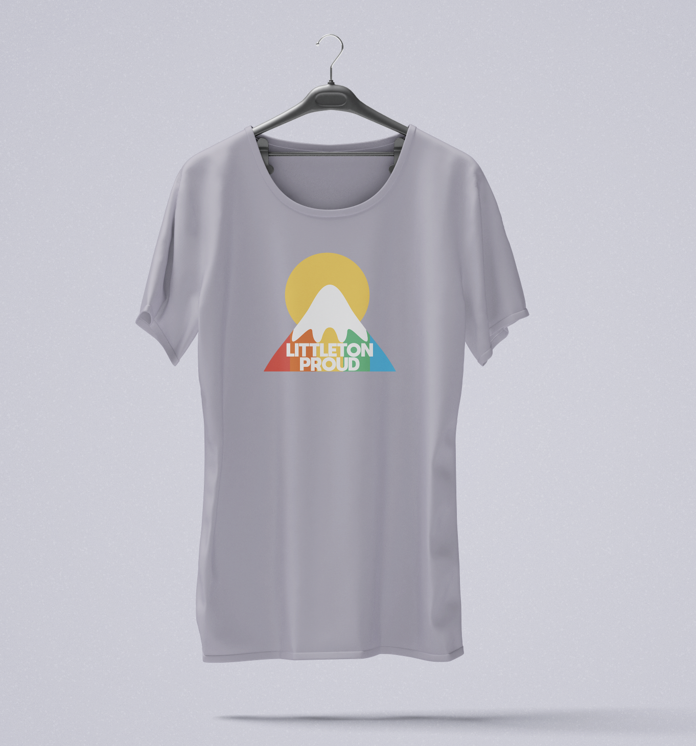 Gray T-shirt hanging on a hanger with a colorful graphic design featuring a mountain, a sun, and the words 'Littleton Proud'.