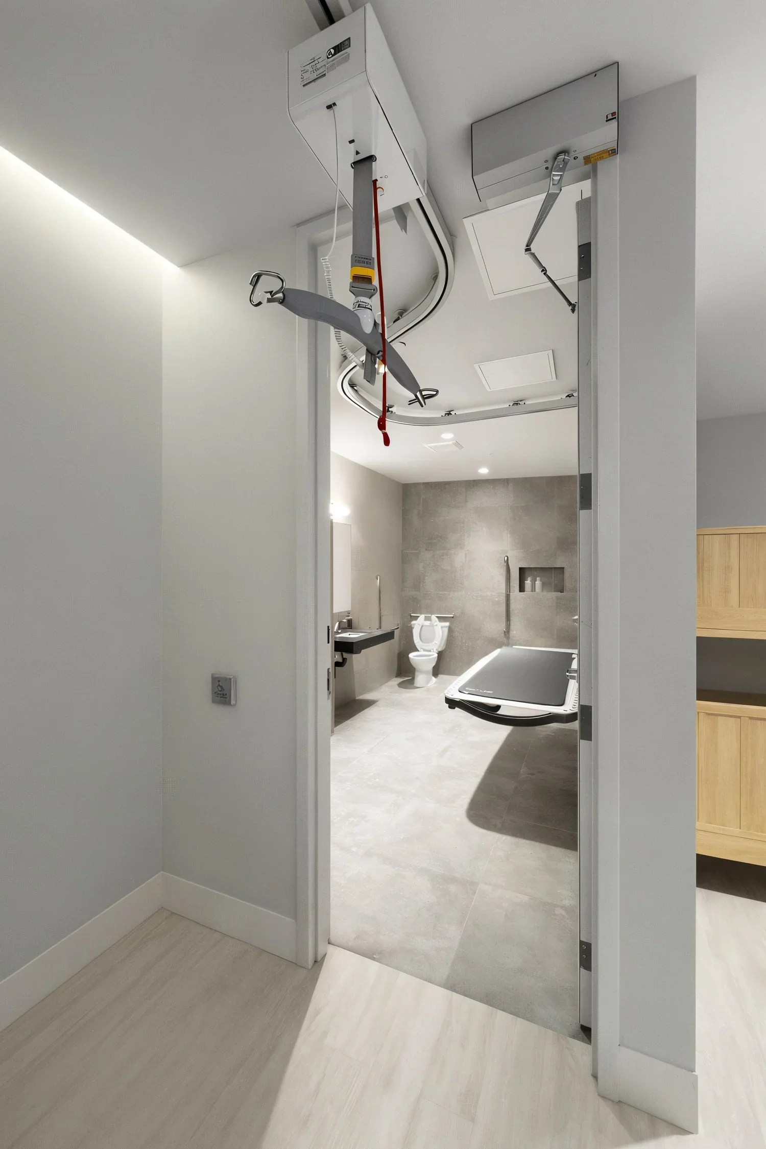 Hospital bathroom with medical equipment including a ceiling lift and a commode, seen through an open door.