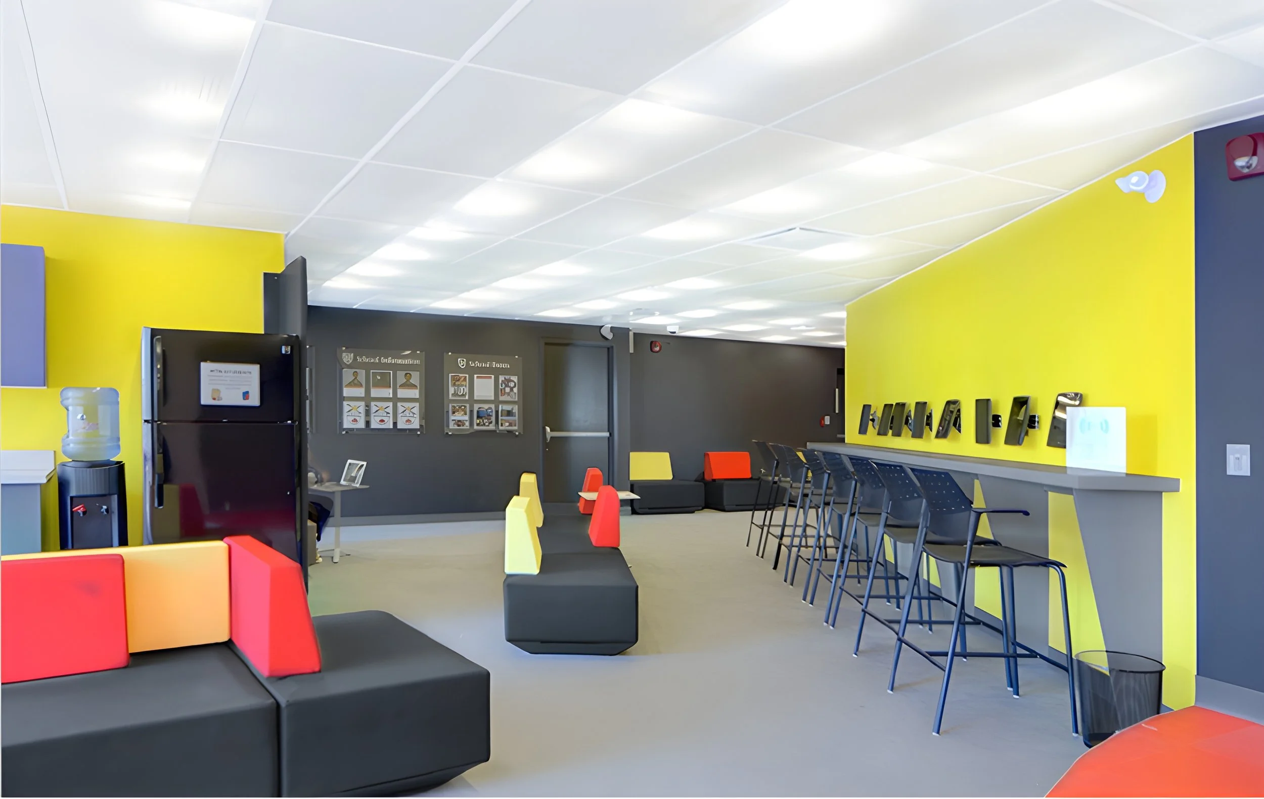 Interior of a modern waiting room or lounge with black, yellow, and gray walls, colorful seating, and a row of black chairs with blue legs. There is a water cooler on the left, framed informational posters on the back wall, and a counter with black a