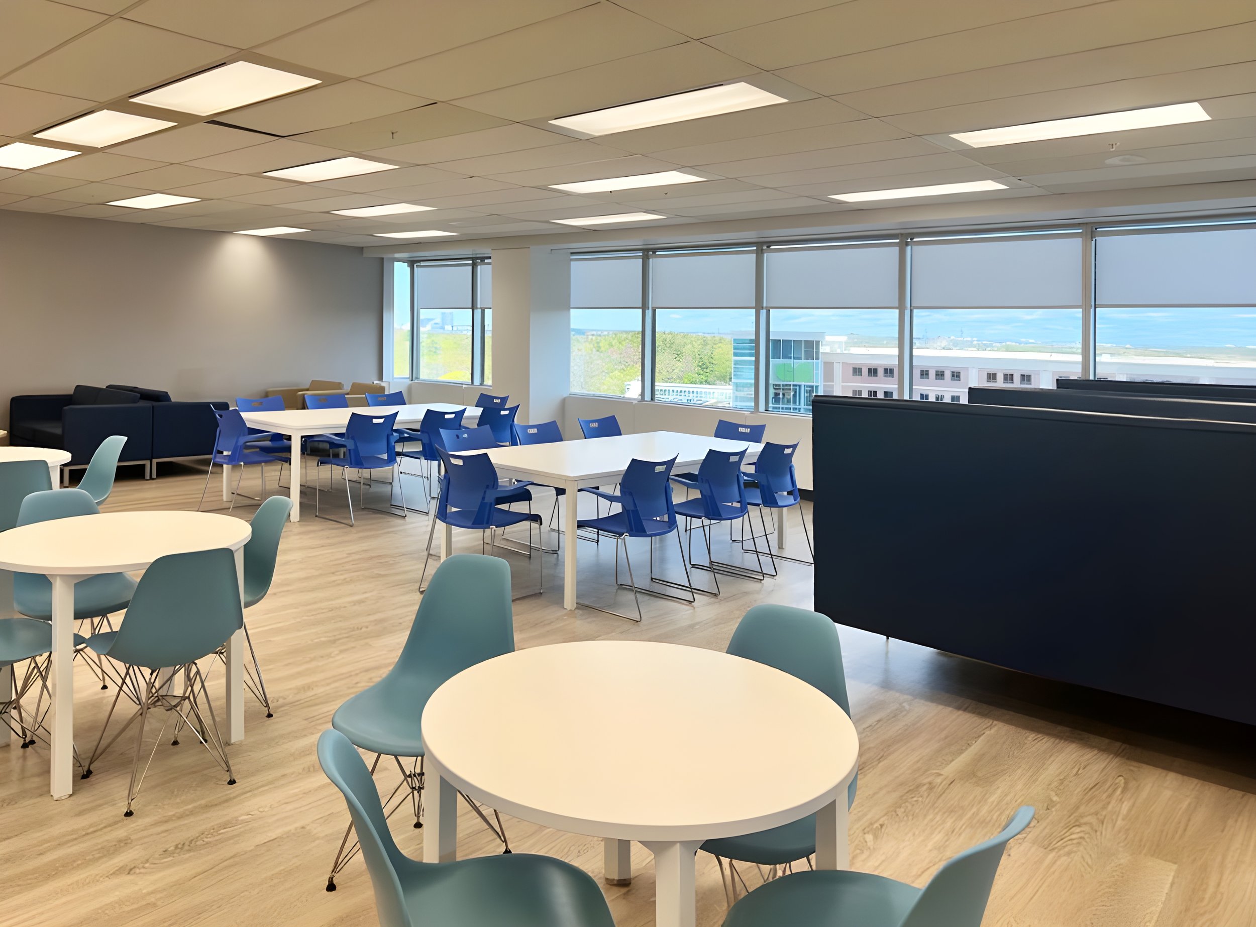 Modern office or conference room with white circular tables, light blue and beige chairs, large windows with city view, and a ceiling with rectangular fluorescent lighting.